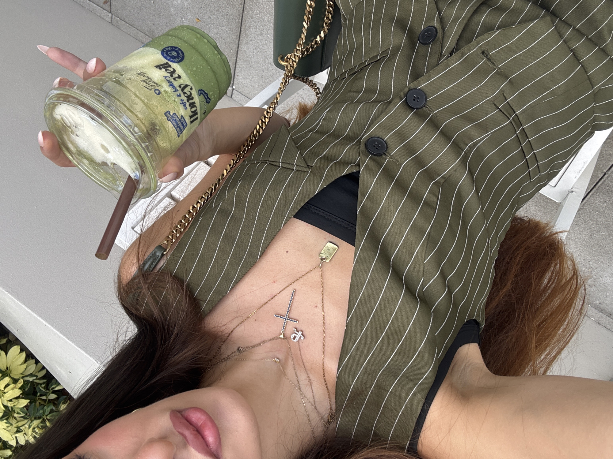 In Hallandale Beach, United States of America, a woman is seen from the chest up, holding a clear plastic cup with a green beverage and a brown straw. The cup has a white lid and features blue and green text that reads "cafe & bakery Honey Veil". She is wearing a green pinstriped vest over a black garment, and multiple gold necklaces adorned with a cross and a small rectangular tag. The background includes a white structure, green foliage, and a portion of a dark green purse with a gold chain strap.