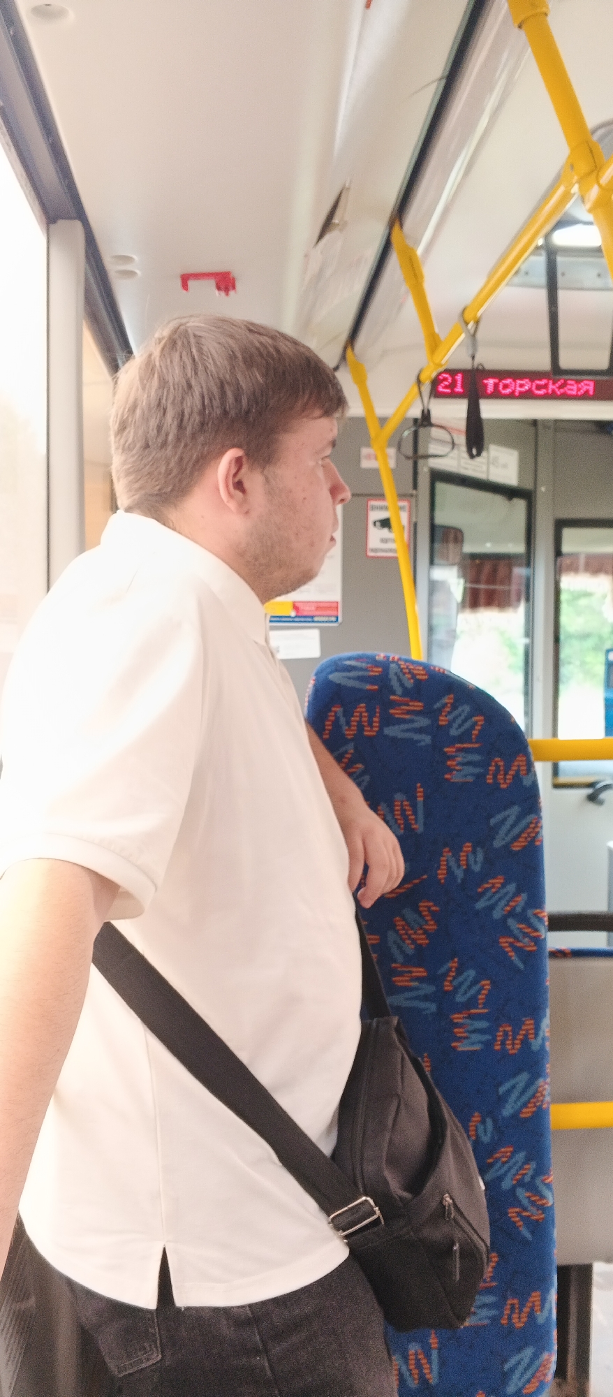 A man with light brown hair and a light complexion is standing inside a public bus. He is wearing a white polo shirt and dark pants, with a black messenger bag slung across his chest. The man is facing to the right, looking out of frame. The bus interior is visible, with yellow handrails, blue and orange patterned seats, and a digital display showing "21 ТОРСКАЯ" which likely indicates the bus route number and destination. The scene appears to be from Izhevsk, Russia, based on the Cyrillic text on the display.