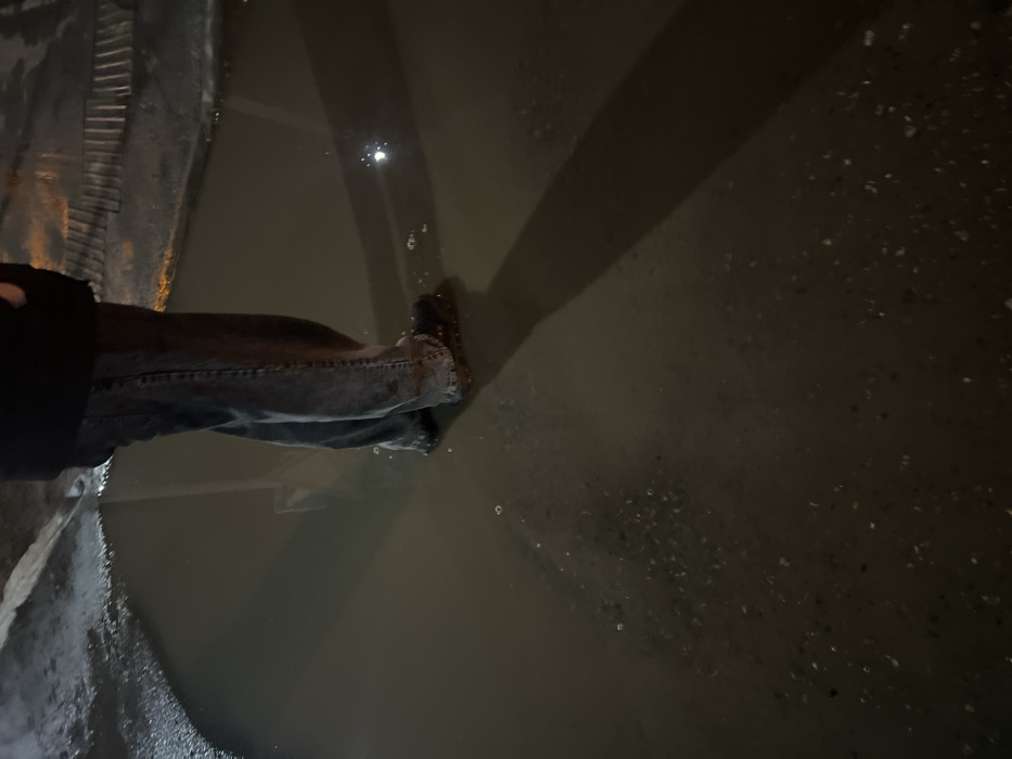 An individual is depicted standing with both feet submerged in a large, murky water puddle at night in Yelabuga, Russia. The person wears dark-colored jeans, dark footwear (likely shoes or boots), and the lower portion of a dark jacket is visible. The water is dark brown/grey, with small bubbles present near the footwear. A long shadow extends from the individual's legs across the water. Reflections of external light sources are visible on the water's surface, with one prominent circular reflection in the lower right. The ground immediately surrounding the puddle's visible edge appears wet and dark.