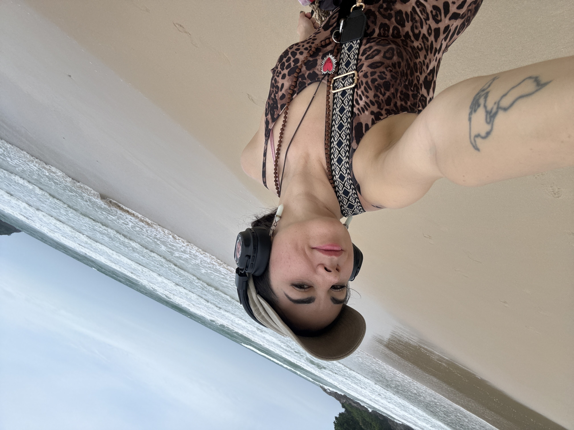 A woman with a leopard print dress and a baseball cap is taking a selfie on a beach in Karon, Thailand. She is wearing headphones and has a tattoo on her arm. The ocean is visible in the background.