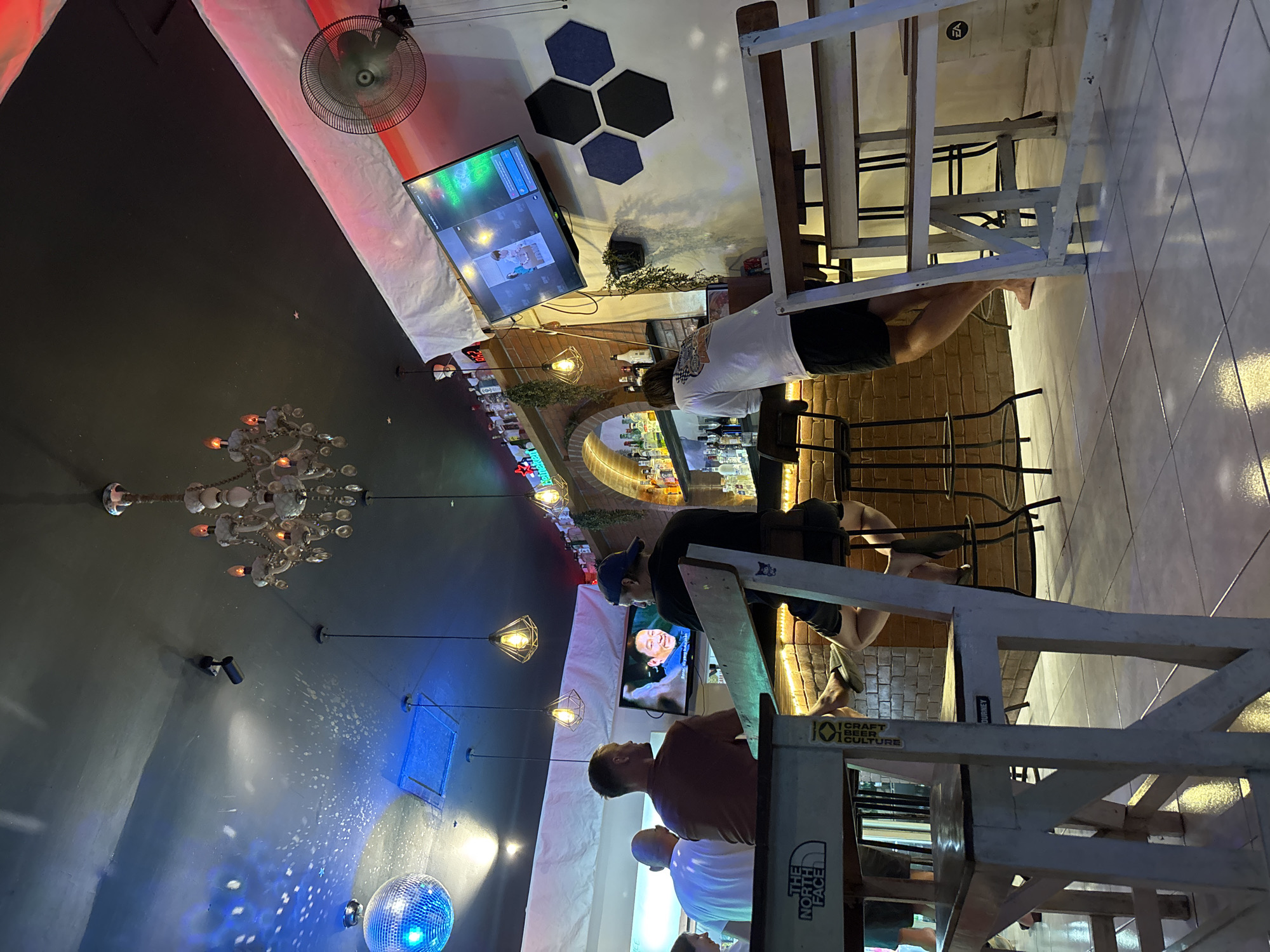 This image depicts an interior scene of a bar, likely located in Kuta Selatan, Indonesia, given the reference location. People are gathered and appear to be seated at the bar. A television is mounted on the wall, displaying content; the context suggests they may be watching something. The bar itself has a selection of bottles. A chandelier and a disco ball add to the ambiance. Furniture like chairs and tables are visible, as well as architectural details like tiled walls. A ceiling fan is also present. The environment seems to be dimly lit and casual. The angle of the shot is unconventional.