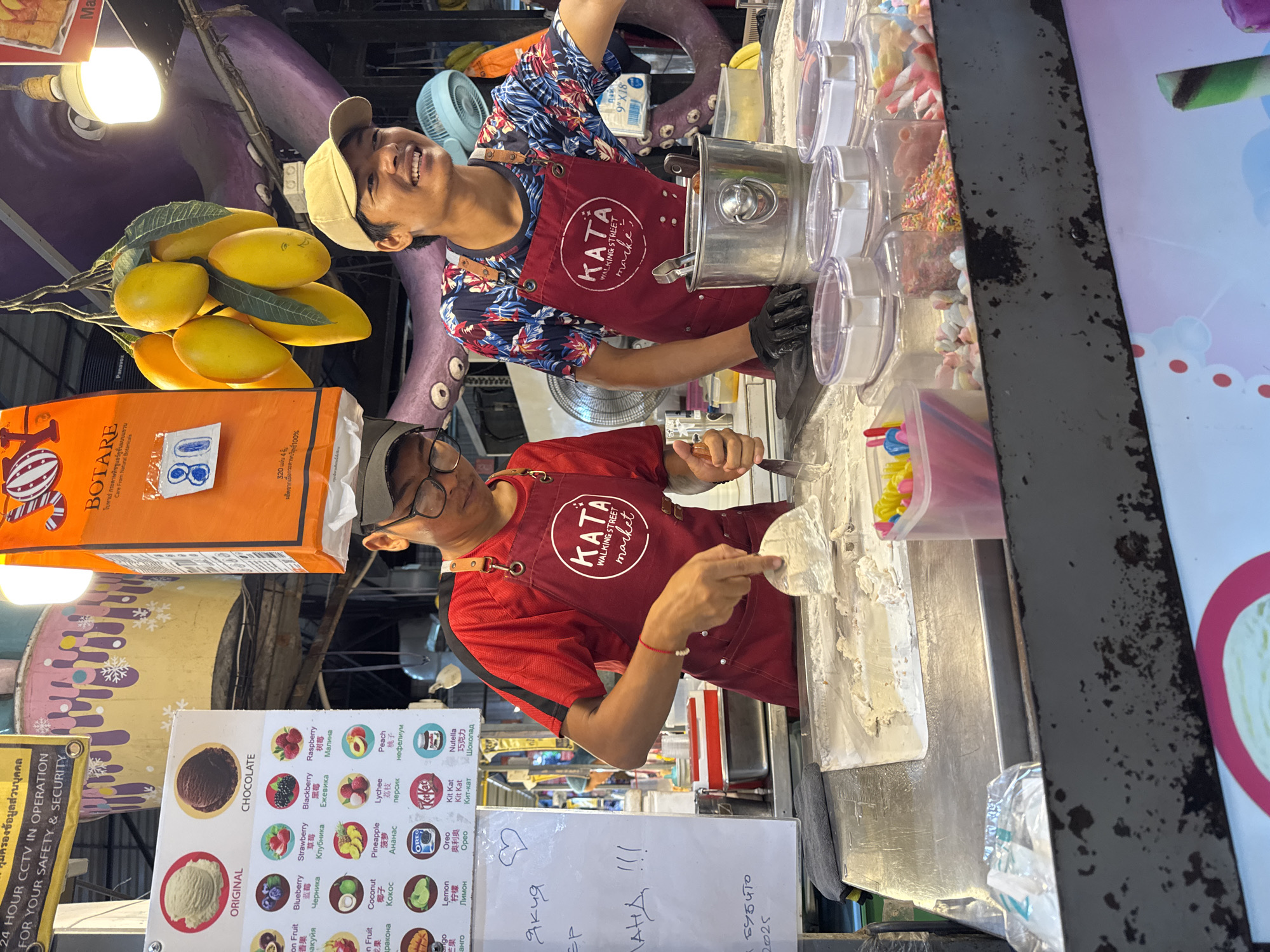 The image depicts a street food stall in Karon, Thailand, where two young men are preparing rolled ice cream. The stall features a stainless steel preparation surface where ice cream is being spread and chopped with spatulas. Various toppings, including sprinkles and marshmallows, are displayed in clear plastic containers. The men are wearing red aprons with the "KATA WALKING STREET MARKET" logo. One man is actively working on the ice cream, while the other is smiling and looking to his right. In the background, colorful decorations and a large, purple octopus-like structure are visible, suggesting a vibrant market atmosphere. A menu board listing various ice cream flavors is also present.