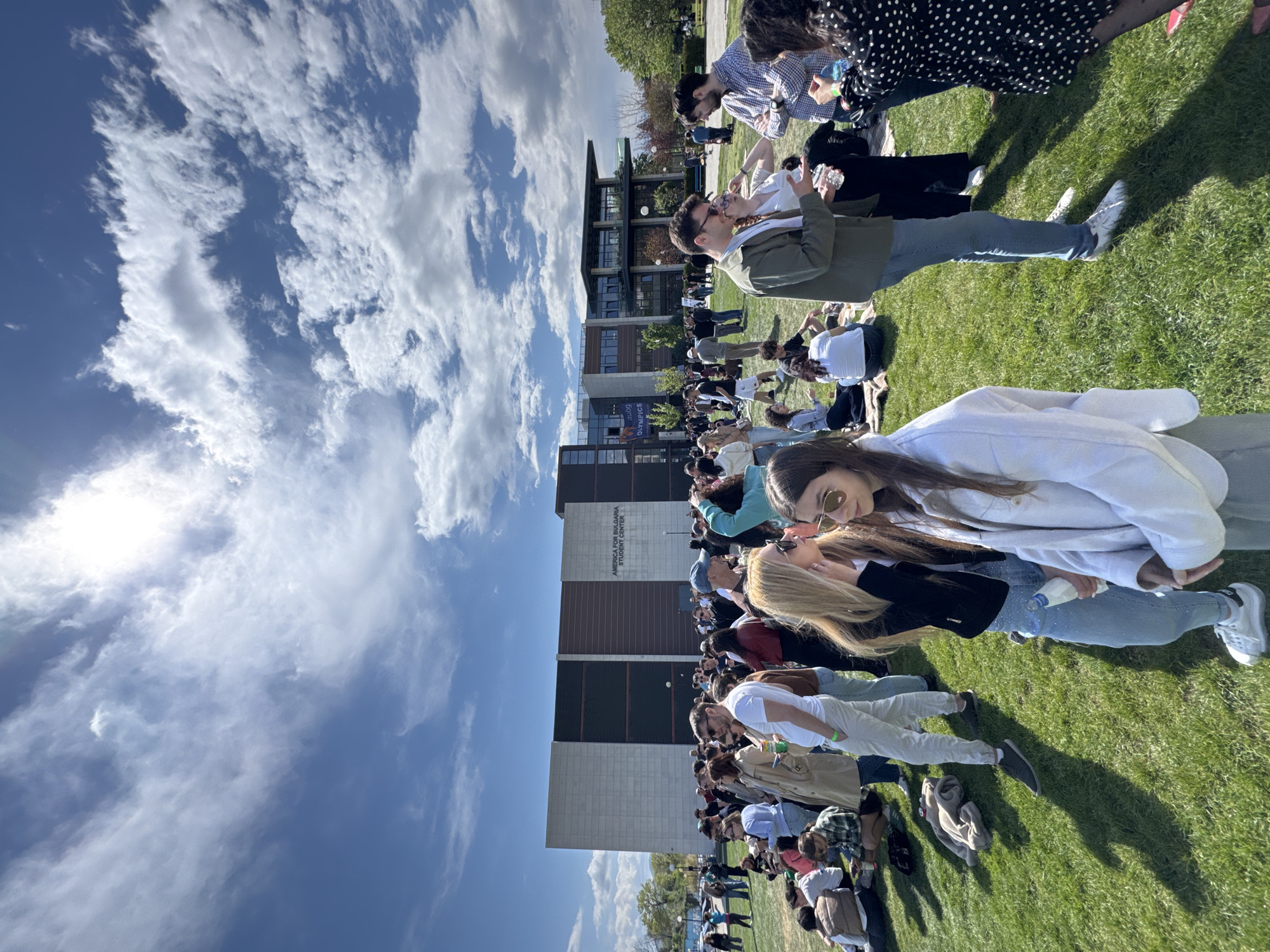 A large group of people are gathered on a grassy area in front of a multi-story building. The building has sections with dark windows and lighter-colored stone or brick facades. Some text is visible on the building, reading "America for Bulgaria Student Center". The sky is blue with scattered white clouds. Most people are standing or sitting on the grass. A few people are closer to the camera, showing details such as clothing and sunglasses. It is daytime, with bright sunlight. The location is in Blagoevgrad, Bulgaria.