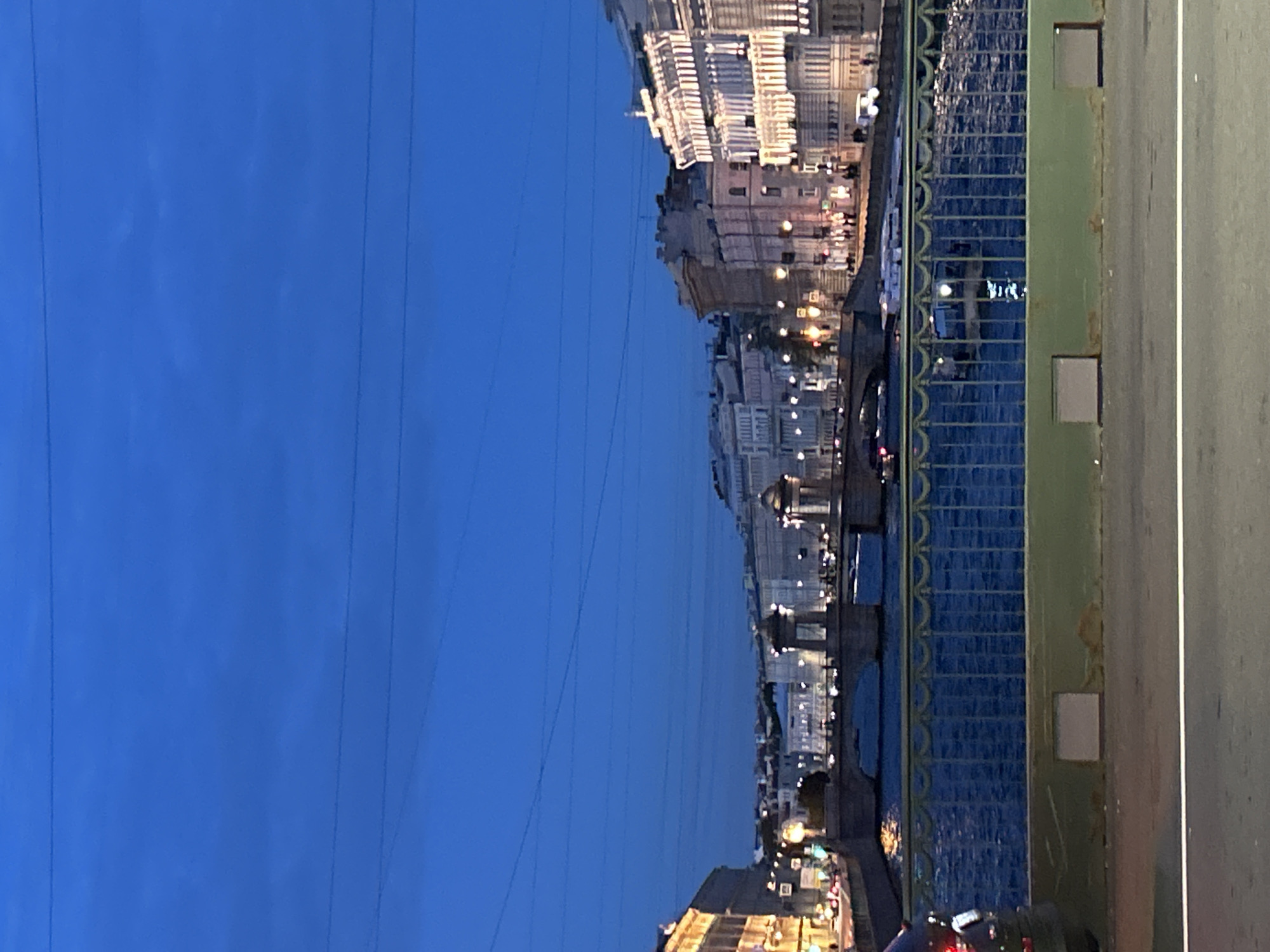 A long shot captures a city skyline at dusk over a wide river. The sky is a deep blue, with thin, dark power lines stretching across it. Buildings line the riverbanks, illuminated by warm lights, their reflections shimmering on the water's surface. A green railing with an ornate pattern runs along the foreground, separating the viewer from the river. A bridge spans the river, its stone arches visible in the distance, with cars and pedestrians crossing it. Boats are visible on the water, their lights adding to the city's glow. The overall scene evokes a tranquil, urban atmosphere as day transitions into night.