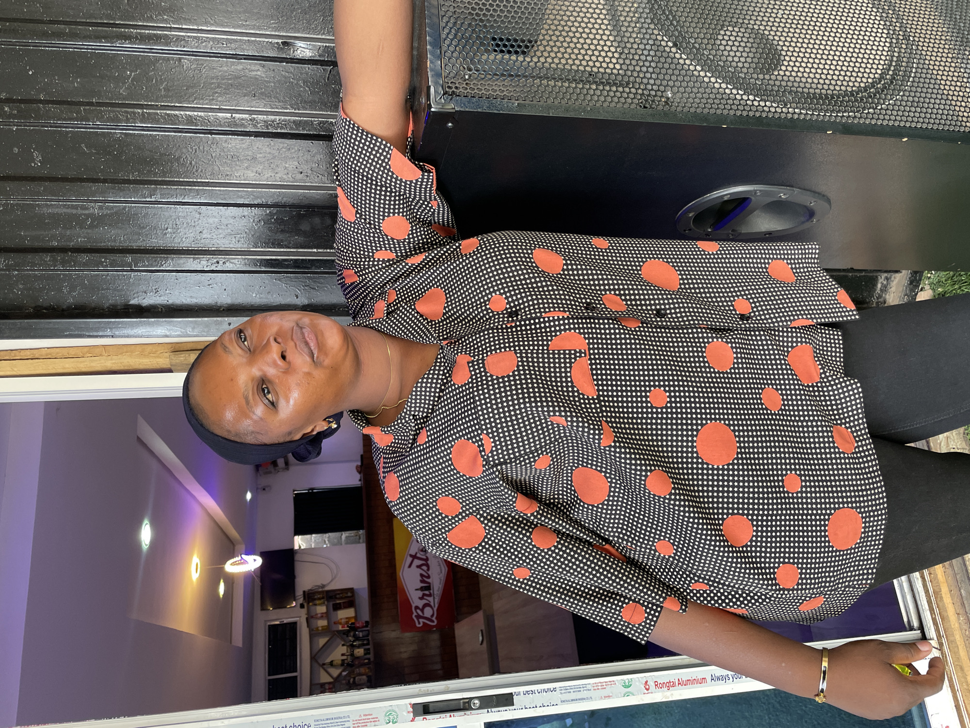 A woman stands in front of a building, possibly a shop or business, in Jos, Nigeria. She is wearing a black and white polka dot shirt with large orange circles, black pants, and a black head covering. She has a gold bracelet on her left wrist and a gold necklace.
She is positioned next to a large black speaker. In the background, through the building's entrance, there is a glimpse of the interior. The interior appears to be a store, with shelves and a "Brinsta" sign visible. The interior lighting includes a mix of white and gold tones. The entryway consists of white aluminum frames and transparent panes.