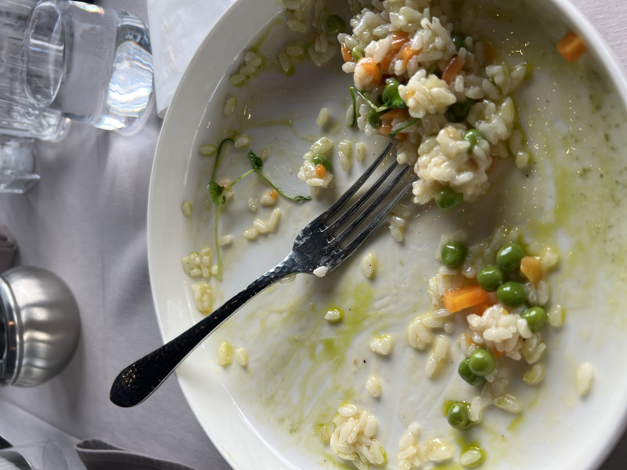 The image shows a mostly eaten plate of risotto with peas and carrots, alongside a fork. The plate rests on a light gray tablecloth. To the left, there is a glass of water and a silver-colored object. No people are visible. The location is unknown, and there is no evidence indicating it is Burgas, Bulgaria.