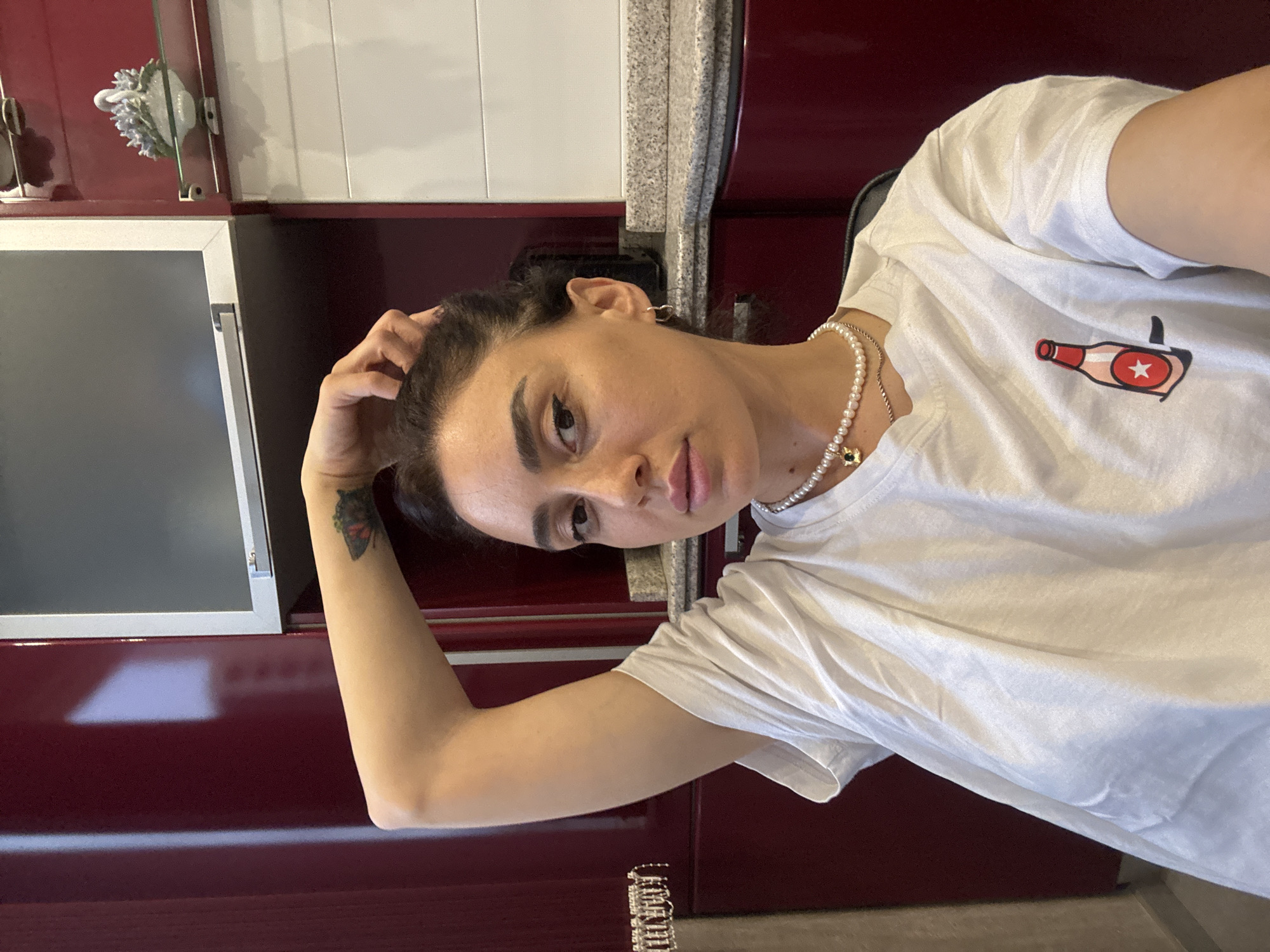 The image shows a woman with dark hair pulled back, wearing a white t-shirt with a graphic of a pink bottle with a red star. She has a pearl necklace and a silver chain. Her right arm is raised, with her hand touching her head. She has a tattoo of a butterfly on her upper arm. The background appears to be a kitchen with dark red cabinets and speckled countertops. A small white swan figurine is visible on a shelf in the background. The lighting suggests an indoor setting, possibly a home. The location is not specified.