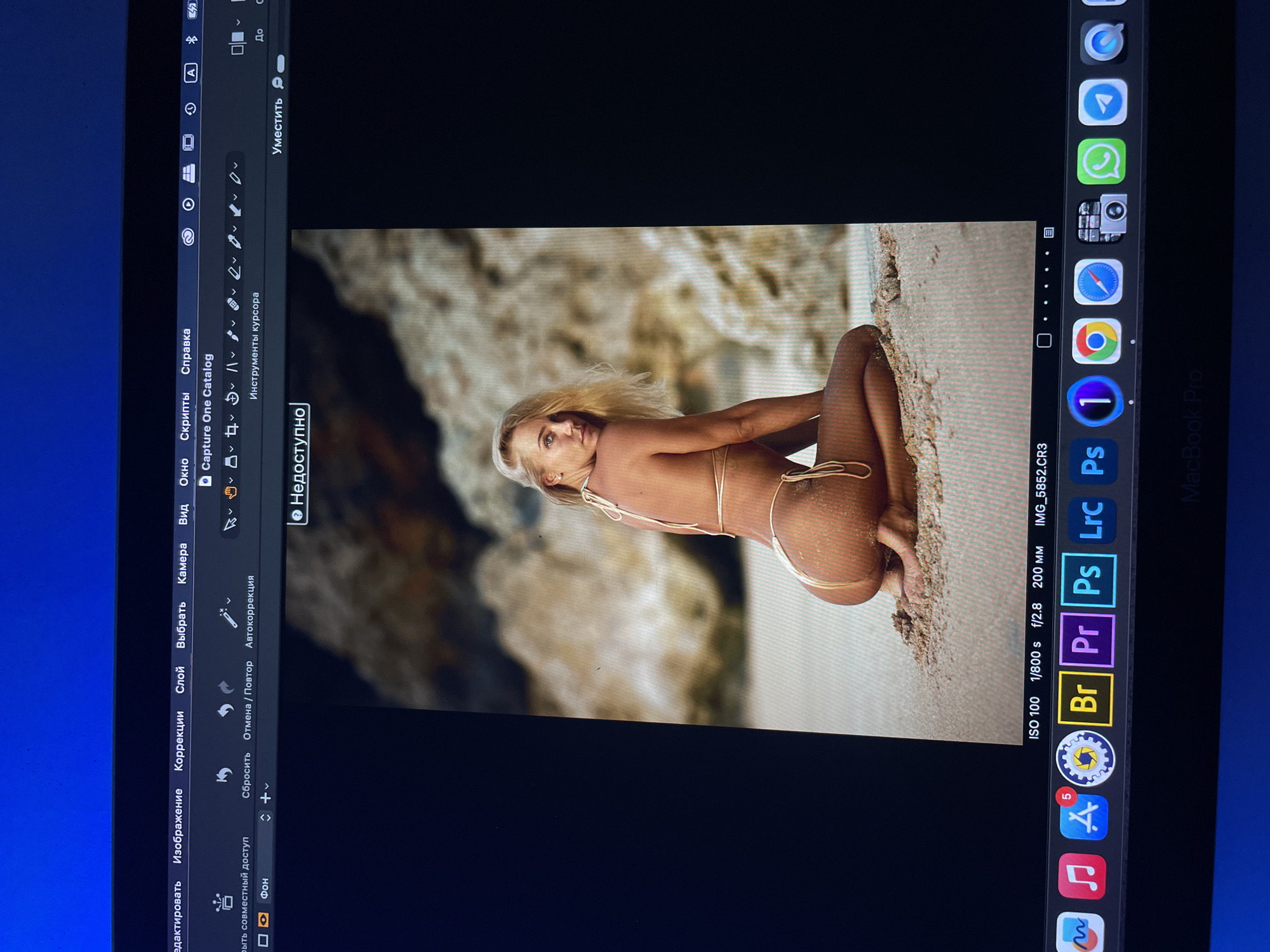 The image shows a computer screen displaying the Capture One photo editing software. The software has an open image of a blonde woman on a beach, wearing a skimpy bikini, appearing to pose for a photograph. The screen also shows other apps, including Adobe Photoshop, Premiere Pro, Lightroom, and Chrome. The computer itself is a MacBook Pro. It is not possible to identify the specific location in the city of Kazan, Russia, from this image.
