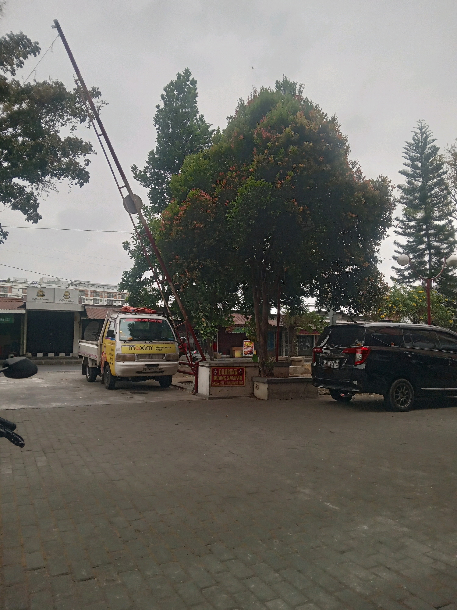 The image depicts a street scene in Depok, Indonesia, likely in a commercial or residential area. In the foreground, a gray paved surface suggests a road or parking area. A red and white boom gate, partially lowered, is a prominent feature. To the left of the boom gate, a yellow and white pickup truck with the "Maxim" logo is parked. The truck has Indonesian license plates and displays promotional text. Behind the truck, a row of businesses with closed shutters are visible, one with signs indicating "Coffee & Eatery." To the right of the boom gate, a black SUV is parked, also with Indonesian license plates.
The background includes lush green trees, one with reddish-tinged leaves, and a tall, conical pine tree. In the distance, several modern buildings with multiple floors are visible, suggesting an urban or suburban setting. The sky is overcast, indicating a cloudy day. A small red and white sign with text in Indonesian is affixed to a white concrete structure near the black SUV. A motorcycle is partially visible in the lower left corner of the image.