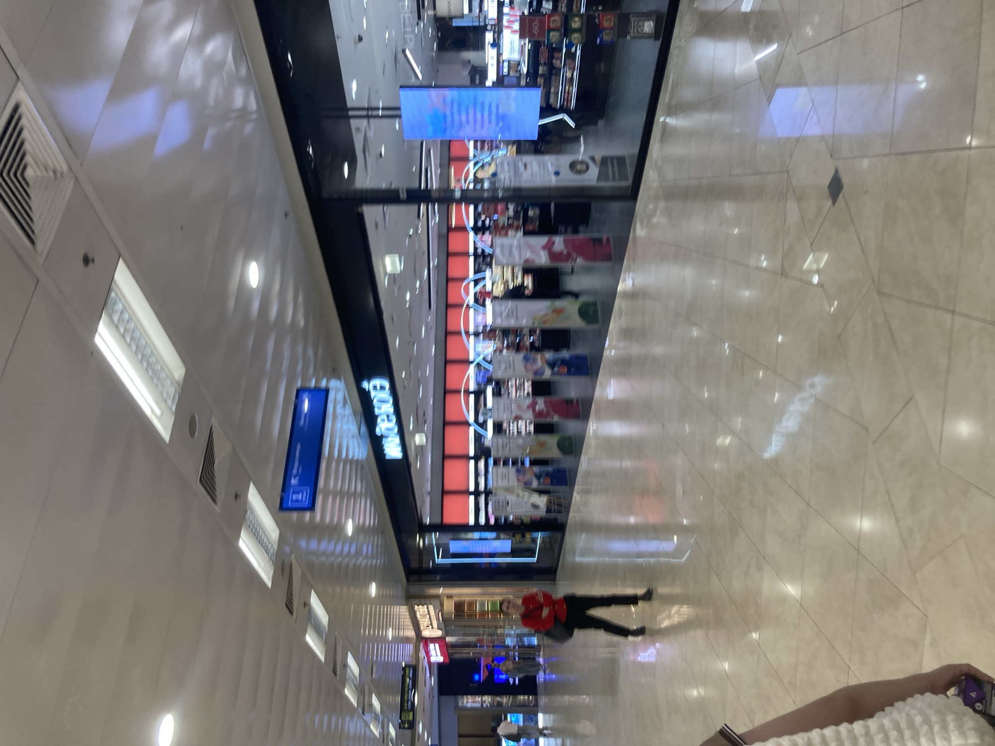 The image presents an indoor view of a modern commercial complex, highly probable to be located in Moscow, Russia, as evidenced by the prominent "ИЛЬ ДЕ БОТЭ" (IL DE BEAUTE) storefront and other Cyrillic signage. The scene is dominated by a wide, well-lit corridor featuring a polished, light-colored tiled floor that reflects the numerous overhead rectangular fluorescent lights, circular downlights, and portions of the adjacent store interiors. The ceiling is composed of white panels interspersed with air vents.
On the right, the large glass facade of an "ИЛЬ ДЕ БОТЭ" cosmetics and perfume store is visible. The store's interior is brightly illuminated with red-accented displays and multiple product stands. A tall, vertical digital display screen inside the store shows a blue and white abstract pattern. Overhead, a blue directional sign featuring an 'i' icon reads "Информация" (Information) and "Место встречи" (Meeting point).
To the left, another storefront with the partial name "ЗНАКОМСТВО" (ZNAKOMSTVO) on its sign is visible further down the corridor.
Three individuals are present. In the mid-ground, a person wearing a red long-sleeved top and black pants, carrying a black shoulder bag, walks towards the viewer. Further down the corridor on the left, two additional individuals are walking away from the camera. In the immediate bottom-left foreground, the left arm and part of the torso of a person wearing a white, textured garment are visible, holding a small purple object. There are no direct interactions observed between the individuals.