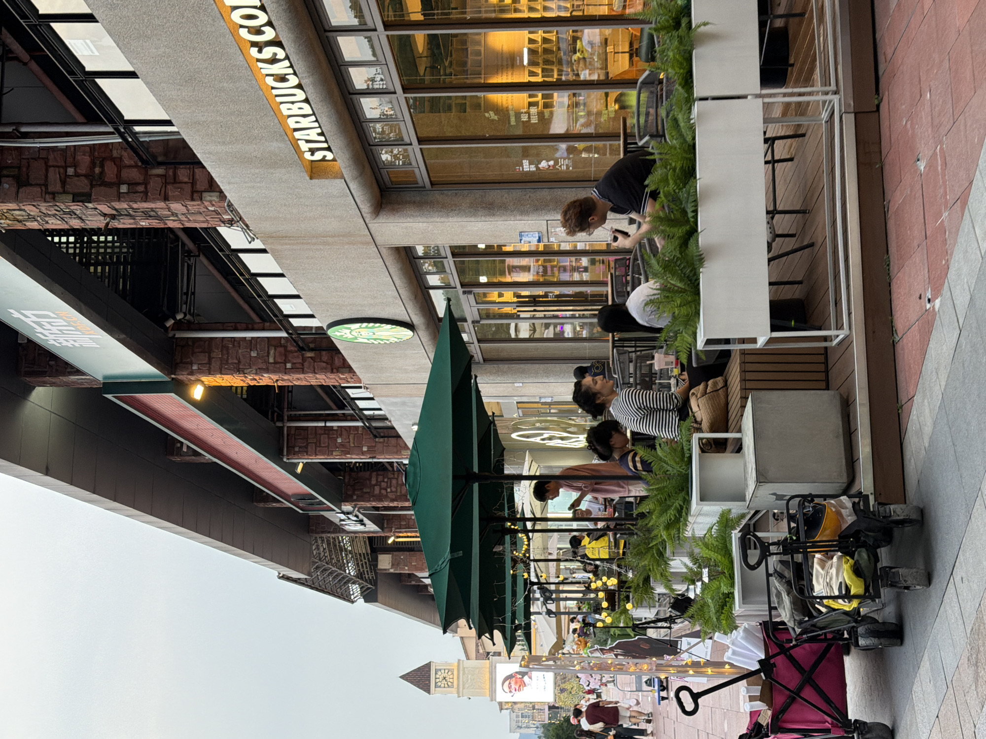 The image depicts an outdoor seating area of a Starbucks store located in the Chongqing High-tech Zone, China. Several people are seated at tables under green umbrellas, engaged in conversations and enjoying beverages. The store facade is visible, featuring the Starbucks logo and signage. In the background, a clock tower stands tall, and other businesses and pedestrians are present, indicating a bustling urban environment. Two red and black carts are placed in the foreground. The overall scene suggests a commercial area with outdoor dining and pedestrian activity.