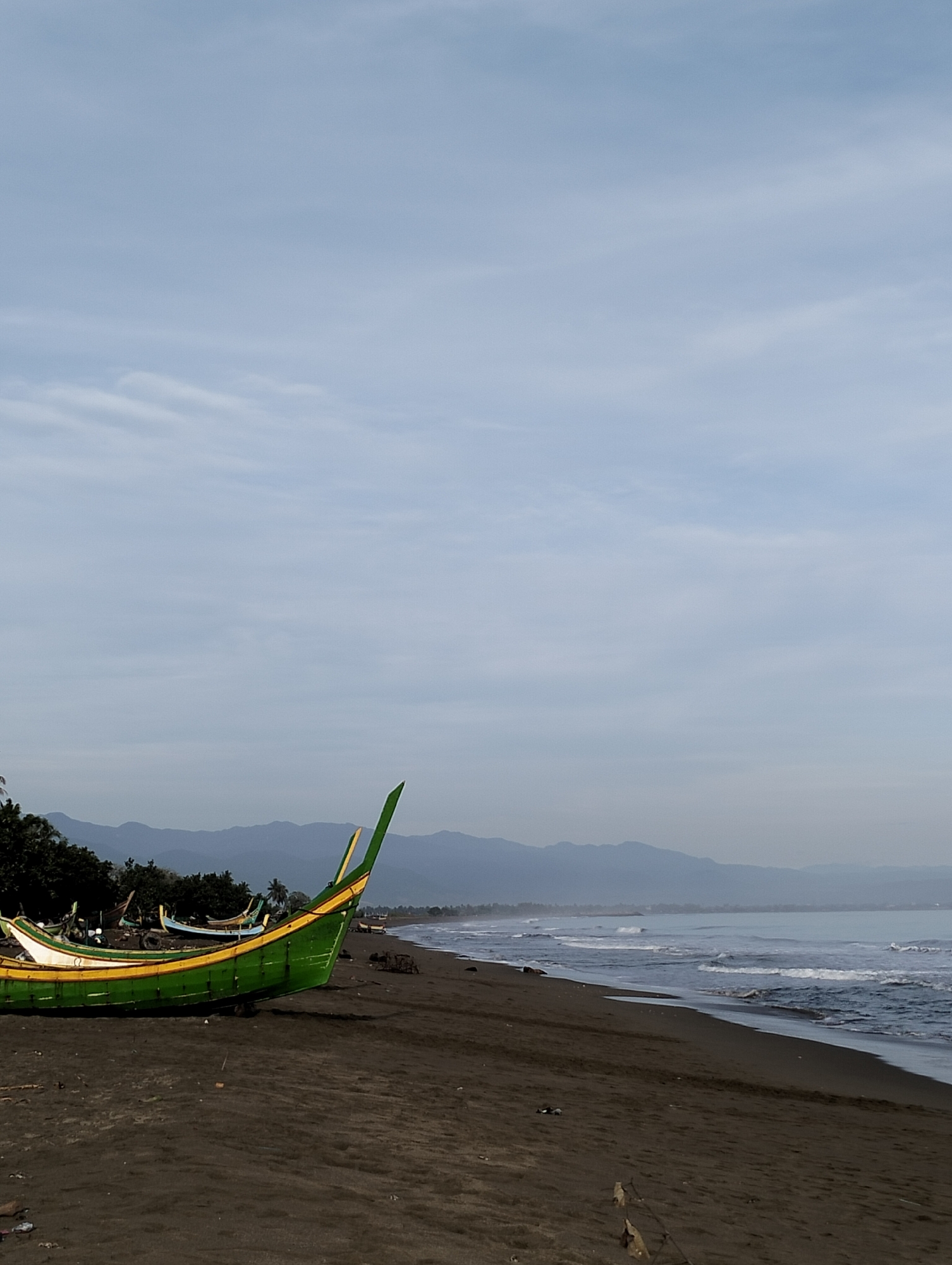 A panoramic view of a coastal scene in Curehtunong, Indonesia, depicts a dark sand beach stretching along a body of water under a light blue, subtly clouded sky. In the foreground, positioned on the left, a large wooden fishing boat, primarily green with prominent yellow and white trim, rests beached on the sand. Several other smaller, less distinctly colored fishing boats are visible further back along the shore, near a dense line of tropical trees that separates the beach from a range of hazy, bluish-grey mountains in the distant background. The sea on the right side of the image shows moderate waves breaking on the dark shore. No individuals are present in the frame. The overall lighting suggests either early morning or late afternoon, with soft, diffuse illumination.