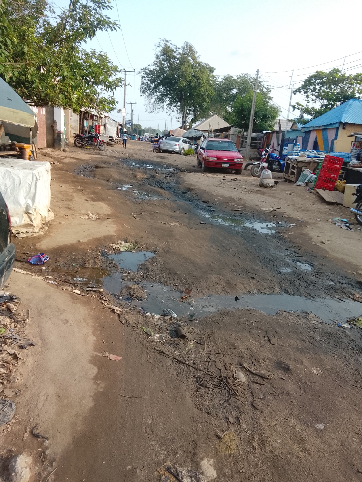 This image depicts a street scene in Abuja, Nigeria. The road is unpaved and appears to be in a state of disrepair, with numerous potholes and muddy puddles. Several vehicles are visible, including a red sedan, a white sedan, and motorcycles. Buildings line the street, some with corrugated iron roofs and others with blue and colorful siding. Vegetation, including trees and bushes, can be seen growing along the sides of the road. There are also piles of debris and discarded items scattered on the ground. A few individuals are present, with one person standing near a motorcycle and another sitting on the ground near a pile of bags. The sky is clear and bright, suggesting it is daytime.