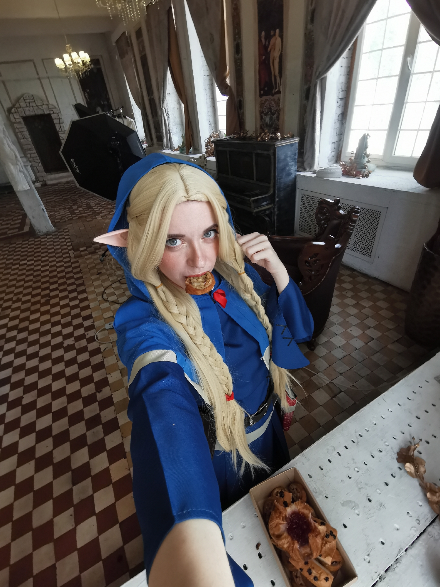 The image depicts a person in cosplay, likely in Moscow, Russia. The individual is wearing a blue hooded costume with long blonde braids adorned with red ribbons, and pointed elf-like ears. They are holding a pastry in their mouth, suggesting they are eating.
The background features a room with tiled flooring, tall windows draped with heavy curtains, an ornate piano, and what appears to be photography equipment. The style of the room suggests a historical building, potentially a museum or a rented studio space. In the bottom right corner, a table is visible with a box of pastries on it.