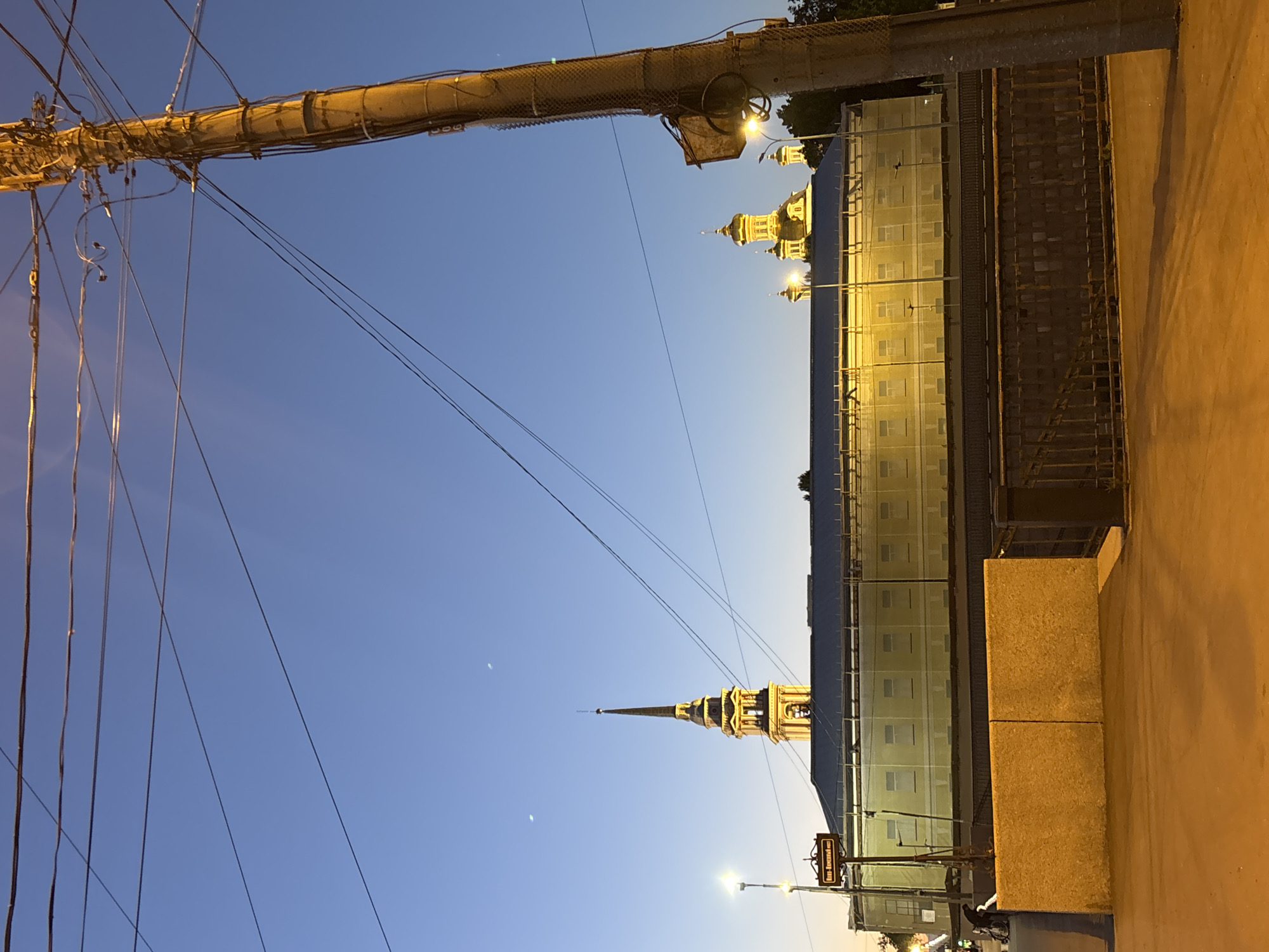 The image depicts an urban scene at dusk in Saint Petersburg, Russia. A tall, slender church spire, illuminated from within, dominates the center-left of the frame, rising above a long, pale yellow building with numerous windows. To the right of the spire, several golden domes of another church are visible. The sky is a gradient of deep blue at the top transitioning to a lighter hue near the horizon. Multiple power lines crisscross the sky, and a large, weathered utility pole with numerous wires and a small basket attached is prominent on the right side of the image. In the foreground, a textured concrete wall runs horizontally, separating the viewer from a lower level, possibly a walkway or street. A street lamp is visible to the left of the spire, and a street sign with Russian text ("Никольский мост" which translates to "Nikolsky Bridge") is also present. A bicycle and a person can be seen on the left side, near the pale yellow building. The overall lighting suggests it is twilight or early evening, with artificial lights illuminating the buildings and pole.