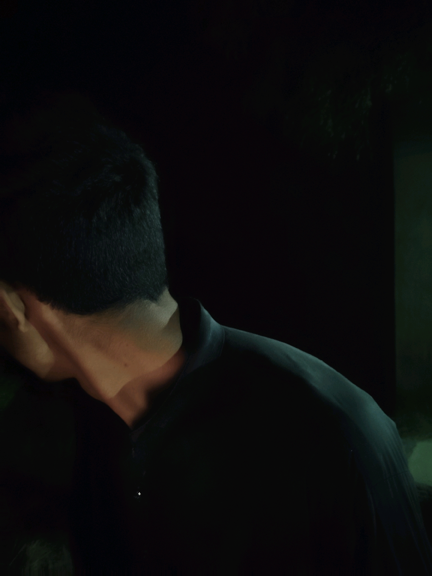 The image shows the back of a person's head and neck, illuminated by a warm light source from the left. The person is wearing a dark shirt with a collar. The background is dark, with some faint greenery visible on the right. The scene appears to be outdoors at night. The image does not provide any specific details about the city of Kot Addu, Pakistan.