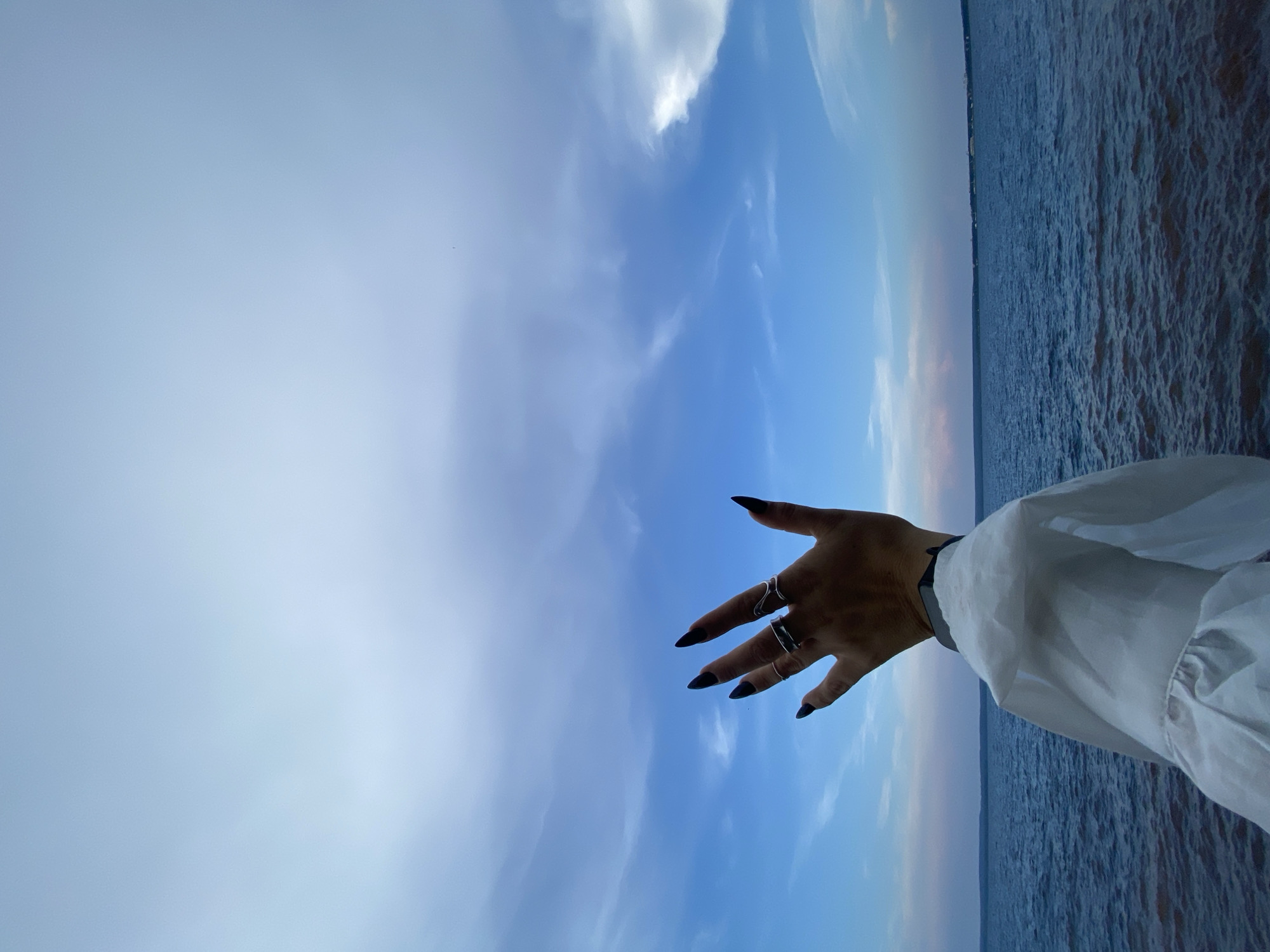 The image shows a person's left hand extended upwards with fingernails painted black and several rings on their fingers. The hand is positioned in front of a cloudy blue sky and a body of water. The person is wearing a white garment with ruffled sleeves. The location is the Kama River embankment in Chaikovsky, Russia.