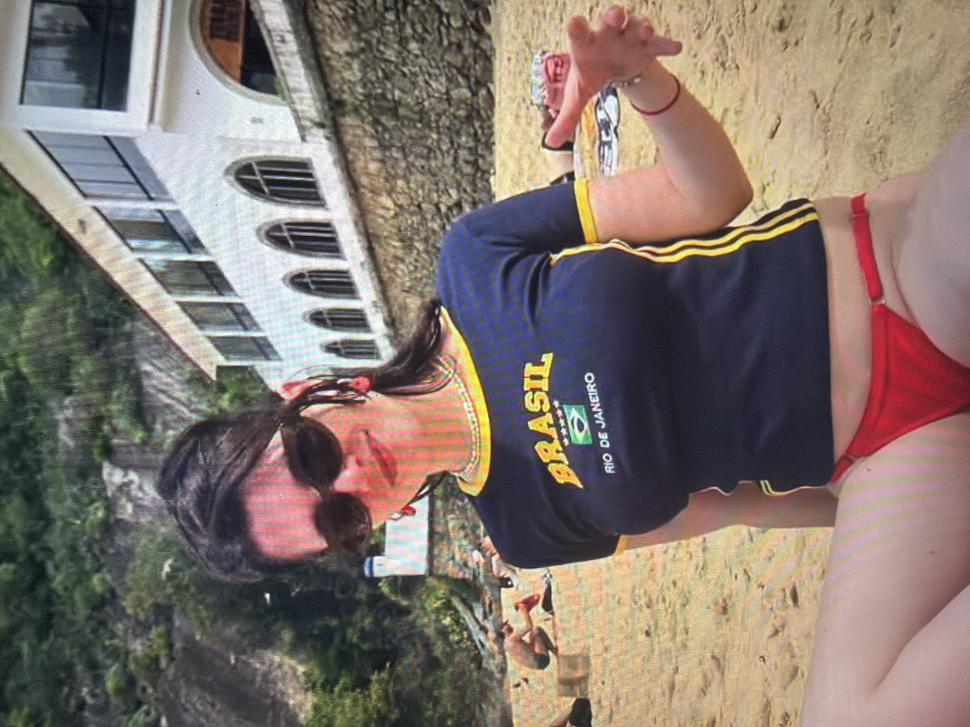 An adult female is depicted seated on a sandy beach, facing forward with a slight smile. She has dark hair, wears dark sunglasses, red drop earrings, and a beaded choker necklace. Her attire consists of a dark blue, short-sleeved t-shirt with yellow trim on the collar and sleeves. The shirt features yellow text "BRASIL" with a Brazilian flag icon below it, and white text "RIO DE JANEIRO." She is also wearing red bikini bottoms. Her left arm is extended towards the viewer, hand open, with a red cord bracelet on her wrist. In the background, a sandy beach extends, populated by several indistinct individuals, one of whom is visible in dark swimwear in a crouched position. Behind the beach, a substantial white multi-story building with numerous windows, including several arched windows on a lower level, stands atop a stone retaining wall. Further in the background, a lush green, tree-covered hillside rises. The scene is brightly lit by daylight. The text on the subject's shirt directly indicates "BRASIL" and "RIO DE JANEIRO" as the depicted location.