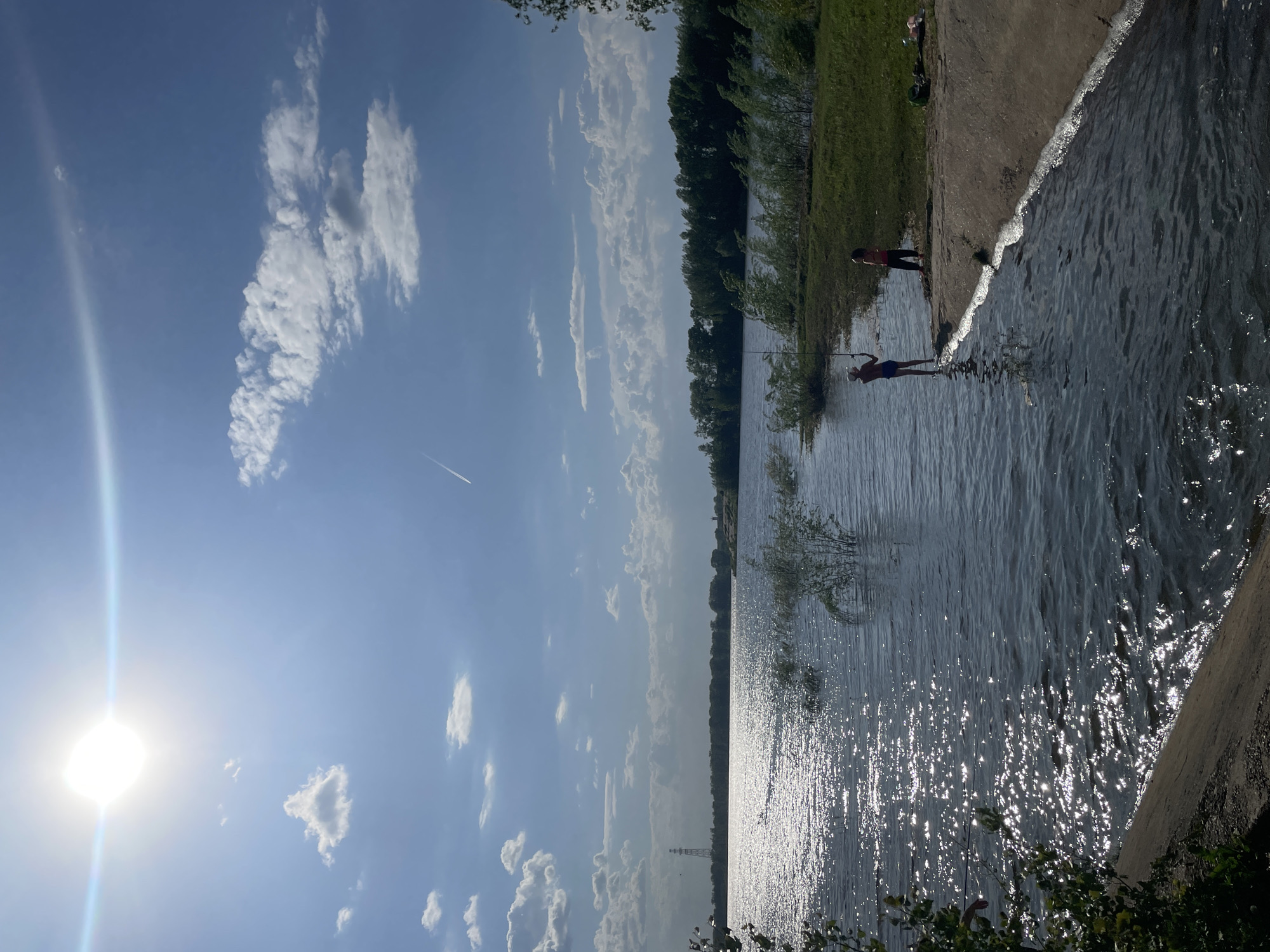 The image depicts an outdoor scene with a body of water, likely a lake or river, extending from the lower right to the middle of the frame. The water surface reflects sunlight, creating bright highlights. The shoreline is visible on the right, with a grassy area behind it. Two people are present near the water's edge; one appears to be wading in the water, while the other stands on the bank. A line of trees runs along the opposite shore in the middle of the frame. The sky above is blue with scattered white clouds and the sun positioned on the lower left. The location is unknown.