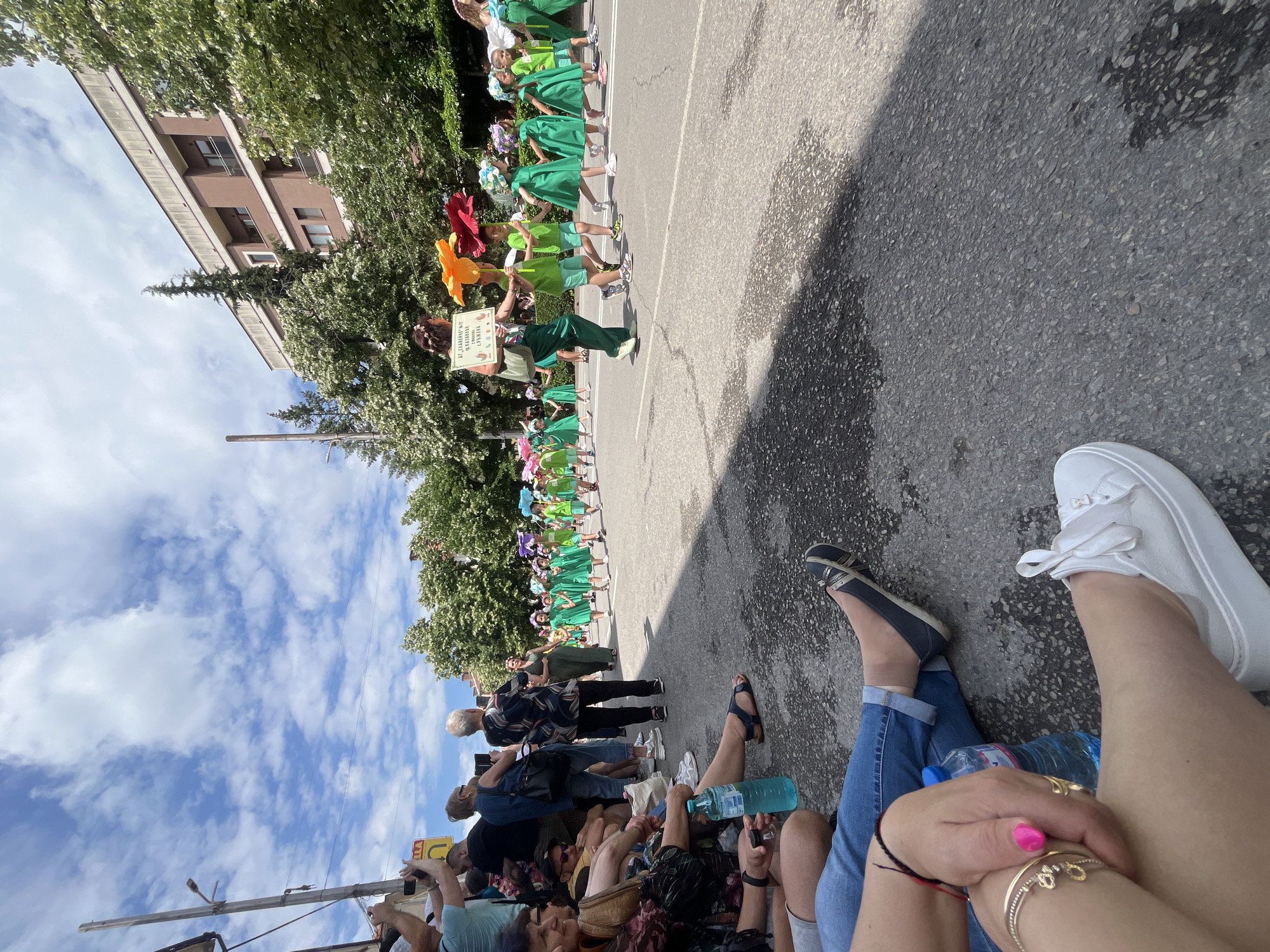 The image depicts an outdoor daytime scene on a city street, likely in Unknown, Bulgaria, due to visible Cyrillic text. In the foreground on the left, several spectators are seated or standing on the pavement, observing a procession. Two sets of legs are prominent: one wearing blue jeans and white platform sneakers with ribbon laces, featuring a gold ankle bracelet and pink toenail polish; another wears blue jeans and dark sandals. A blue plastic water bottle rests on the ground next to them. Further back, more spectators stand, with some holding mobile phones to record the event. A person in a light blue shirt is seen holding a phone aloft, and a woman with grey hair, seen from behind in a patterned top, also holds a phone.
The mid-ground and background feature a children's parade marching down the asphalt street from right to left. Children are predominantly dressed in bright green outfits, with many wearing floral headpieces or carrying large, colorful paper flowers (yellow, red, blue, purple). An adult woman leads a segment of the parade, holding a large yellow paper flower and a white sign with Cyrillic text that translates to "I, the sun and planet Earth. Civil Protection." She is also dressed in green. Another adult woman in a dark green jumpsuit walks behind the children. The parade occupies the width of the street, which shows some dark, wet-looking patches.
The environment includes a clear blue sky with scattered white clouds overhead. Trees with lush green foliage, some exhibiting white blossoms, line the street, providing shade. Multi-story buildings with brown facades, balconies, and windows are visible behind the trees. A street lamp and a wooden utility pole are present on the left side, along with a yellow "U" turn sign that reads "УЛИЦА" (Street) in Cyrillic. The scene is illuminated by strong sunlight, casting shadows on the asphalt.