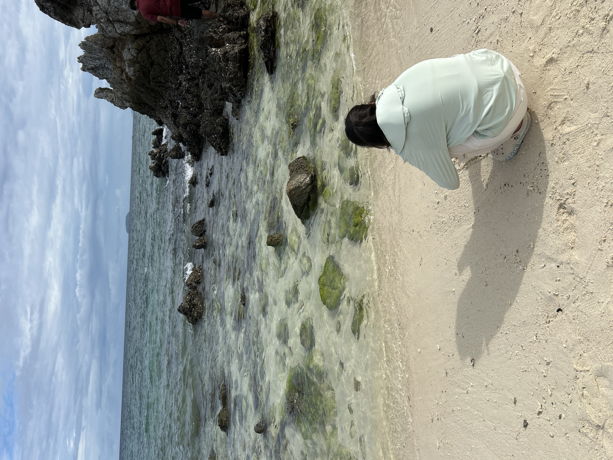 The media file captures a serene beach scene in Tambon Phru Nai, Thailand. The primary subject is a person, viewed from behind, who is crouched down on the sandy shore, seemingly observing the water. They are wearing a light green, long-sleeved shirt and white shorts, with grey slip-on shoes. The person's dark hair is visible.
The setting is a rocky coastline with clear, shallow water lapping at the sandy beach. The water is filled with submerged rocks, some of which are covered in green algae, creating a natural, almost aquatic garden effect. In the distance, the vast ocean stretches out under a sky filled with dramatic, wispy clouds, suggesting it might be late afternoon or a day with changing weather. The sunlight casts a clear shadow of the crouching person onto the sand. To the left of the frame, a large, dark rock formation juts out into the sea. Another person, partially visible and wearing a red shirt and black shorts, stands near this rock formation, also facing towards the water. The overall mood is one of peaceful exploration and appreciation of the natural coastal environment. No visible text is present in the media file.