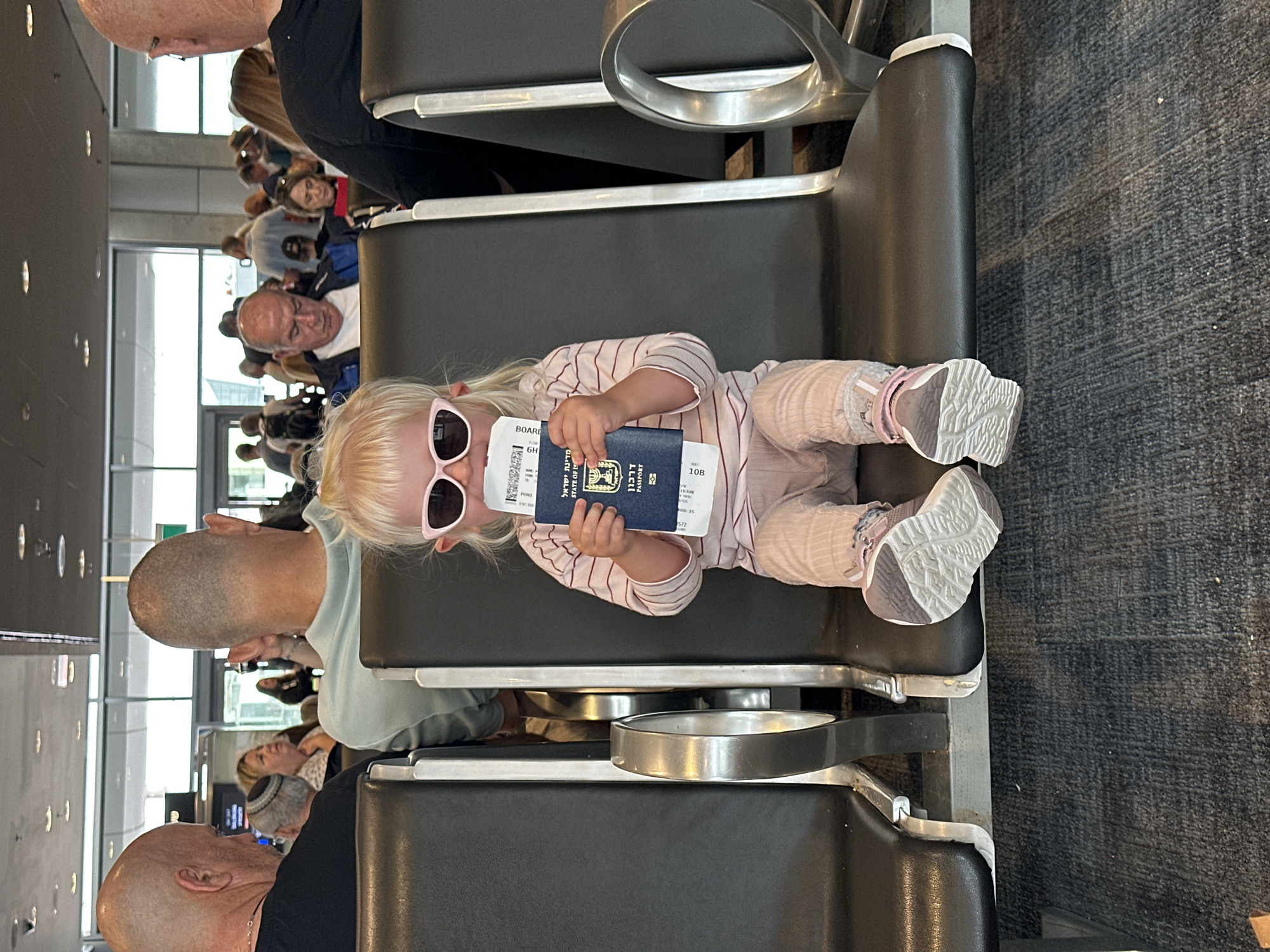 The image shows a young child sitting in an airport waiting area. The child, with blonde hair and wearing sunglasses and a striped shirt, is holding an Israeli passport and boarding pass. The child is seated in a row of black airport chairs. In the background, other travelers are visible, some looking down at their phones or at the departure boards. The environment appears to be a typical airport terminal with a tiled floor and large windows in the distance.