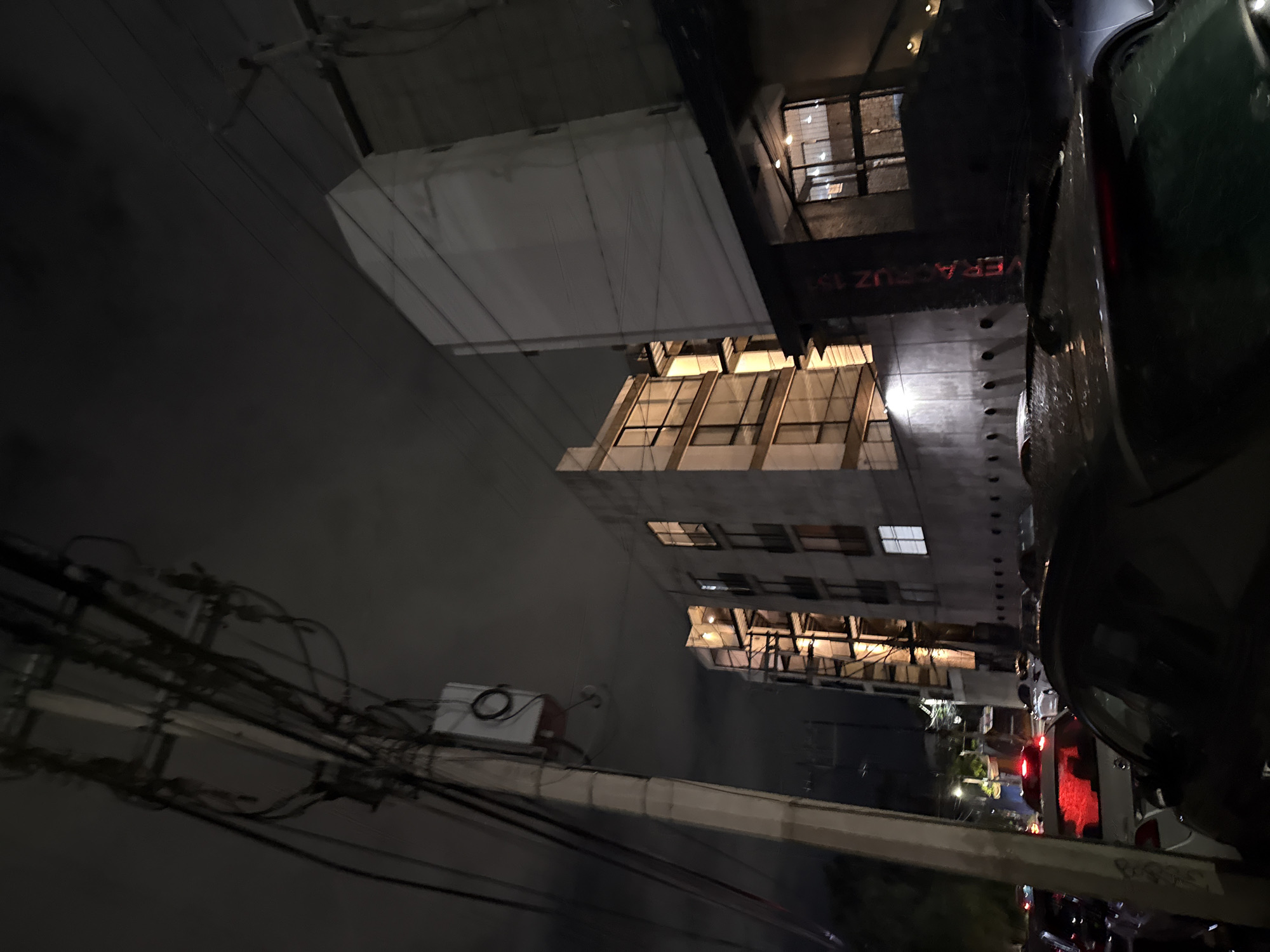 The image depicts a nighttime street scene in Mexico City, Mexico, likely in an urban residential or commercial district. In the foreground, the rear of a dark-colored vehicle is visible, its surface speckled with raindrops. To the left, a utility pole stands tall, adorned with numerous wires and a white electrical box. Behind the pole and vehicle, a multi-story building is illuminated from within, with several windows displaying warm interior lighting. Further to the right, another taller building, with a smooth, light-colored facade, rises into the night sky. Red lettering, appearing to spell "VERACRUZ 13", is visible on a dark column in front of this building. The street appears congested with traffic, with the red taillights of several vehicles visible in the distance. The sky is overcast, contributing to the overall dimness of the scene.