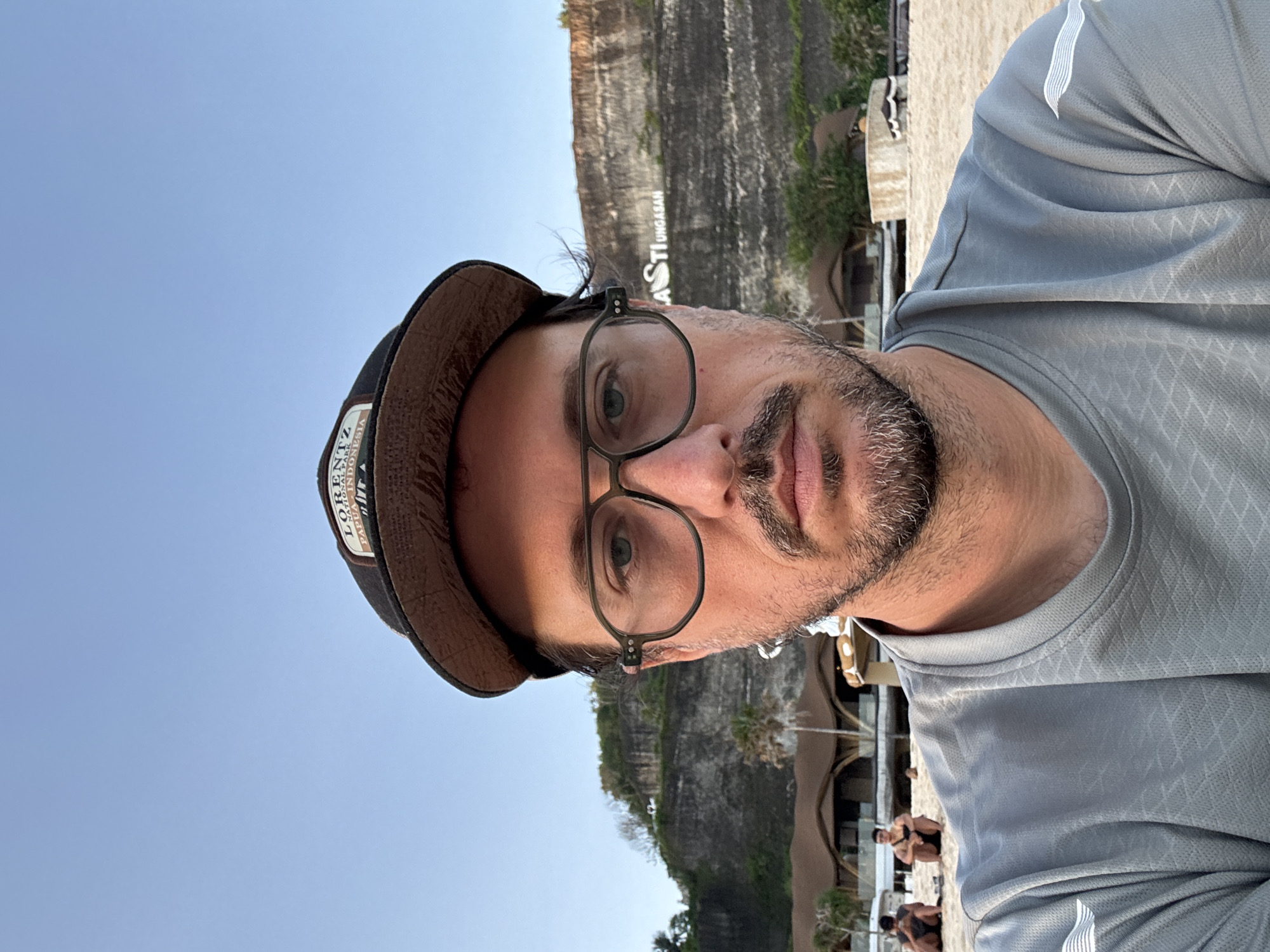 A selfie of a man with glasses and a baseball cap, facing the camera. The man is wearing a gray shirt. The background shows a cliff face with the words "KASTI UNGASAN" in large white letters, indicating the location is possibly near Ungasan in Kuta Selatan, Indonesia. Below the cliff are structures resembling tents or open-air buildings. People are present in the background on the beach. The sky is clear and blue.