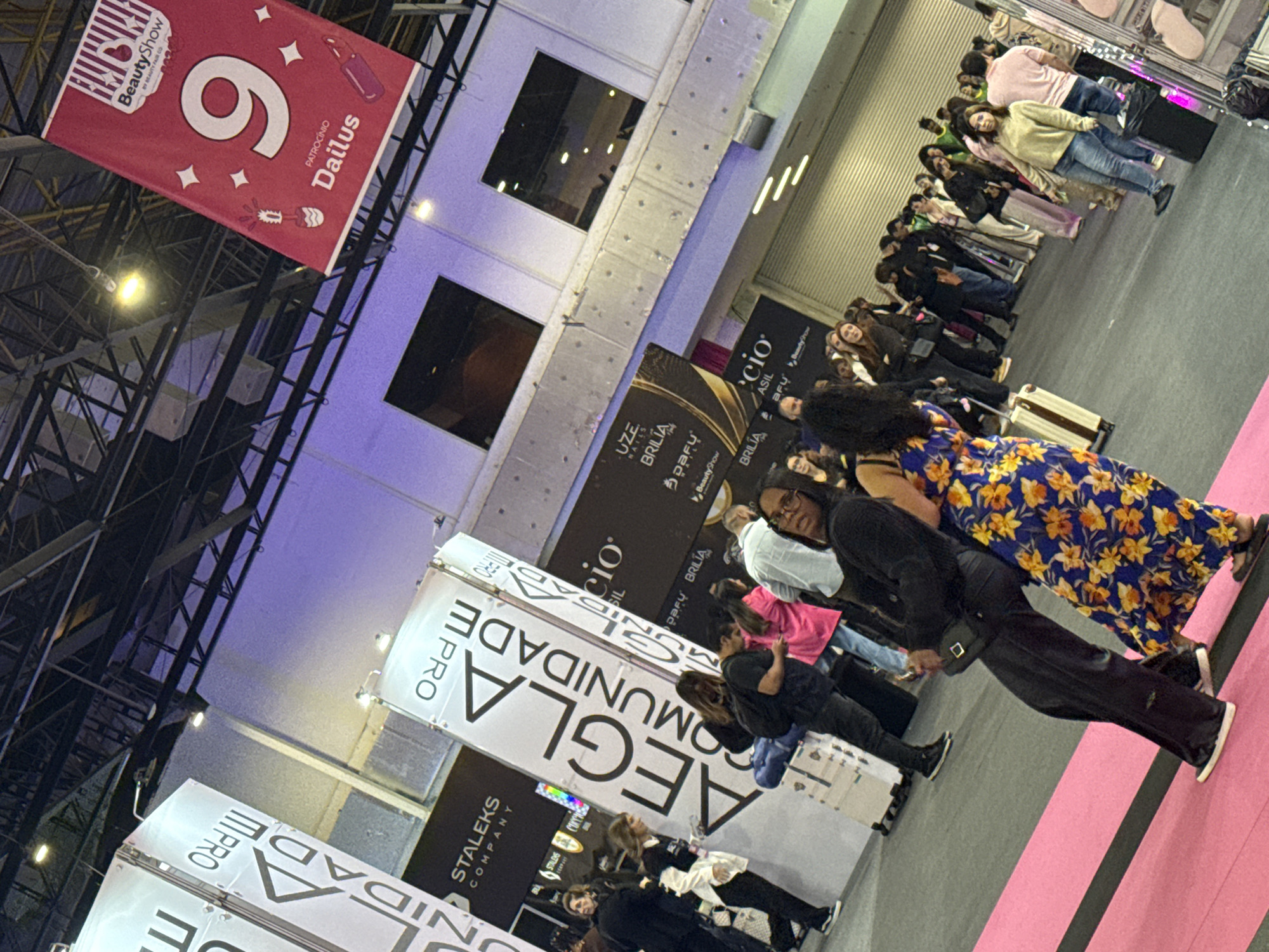 A photograph captures the interior of an event space in São Paulo, Brazil, where a line of people stand along a pink carpet. A banner for the "Beauty Show" is visible, along with advertisements for beauty brands such as Dailus, Brilia, and Stalecks Company. The location appears to be a commercial or trade show environment, with attendees likely participating in or spectating an event related to beauty and personal care.