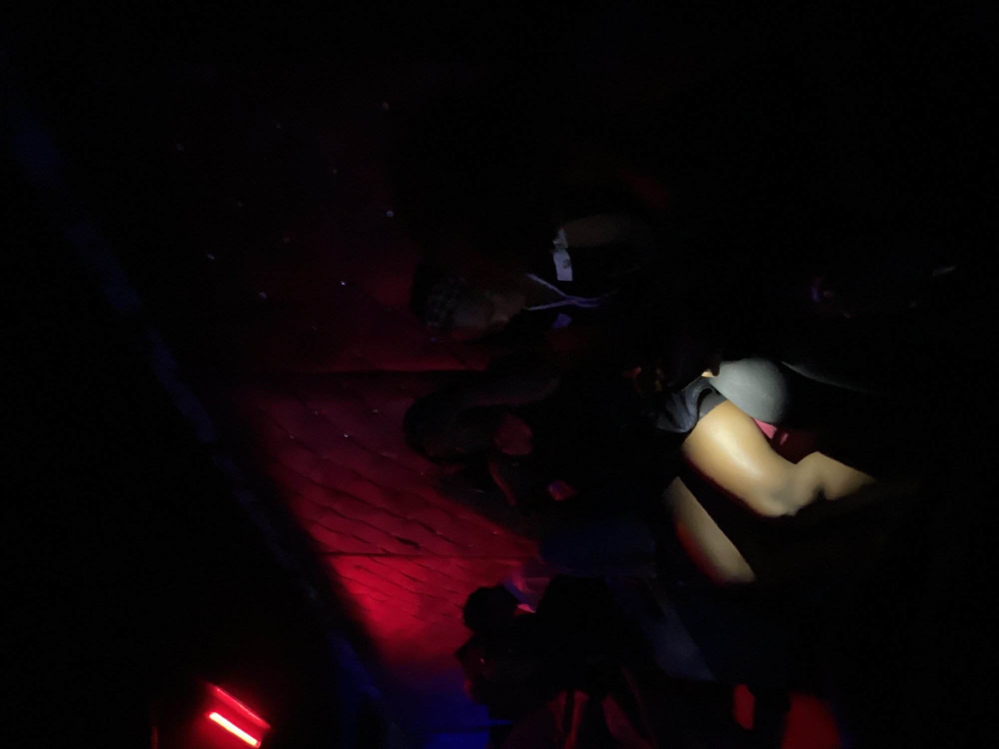 In a dimly lit room, possibly a nightclub or entertainment venue in Durumi, Nigeria, two young women are seated, partially illuminated by red and blue ambient lighting. The woman on the left is positioned closer to the foreground, her legs crossed and her upper body leaning back. Her skin tone is visibly highlighted by the red light, accentuating her knees and thighs. She appears to be wearing a dark top. To her right, the second woman is also seated, facing slightly away from the camera. She has a lighter skin tone and her hair is styled with braids. She is wearing a dark top with white detailing on the sleeve. The background is predominantly dark, with a textured, possibly padded wall that is illuminated by the red light, creating a dramatic effect. The overall atmosphere suggests a lively, possibly intimate, setting.