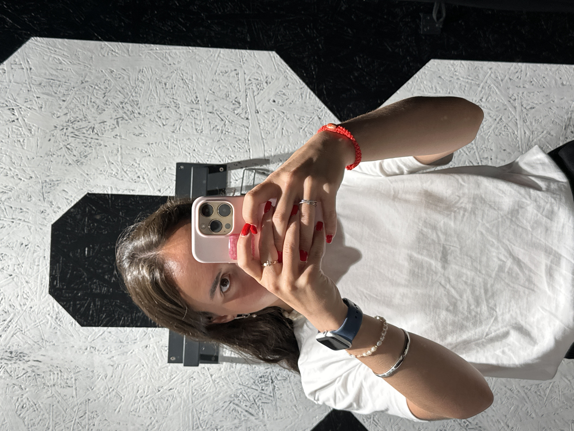A woman is taking a selfie in a room with a distinctive black and white geometric floor pattern resembling interconnected irregular hexagons. The floor appears to be constructed from white wood-chip material painted white and adjacent black hexagonal shapes. The woman is wearing a white t-shirt and holding a pink smartphone with visible camera lenses. She has red nail polish on her fingernails and multiple rings on her fingers. Her left wrist is adorned with a blue smartwatch and two bracelets, while her right wrist has a red woven bracelet. The environment appears indoors. The city of Pattaya City, Thailand is not discernible from the image.