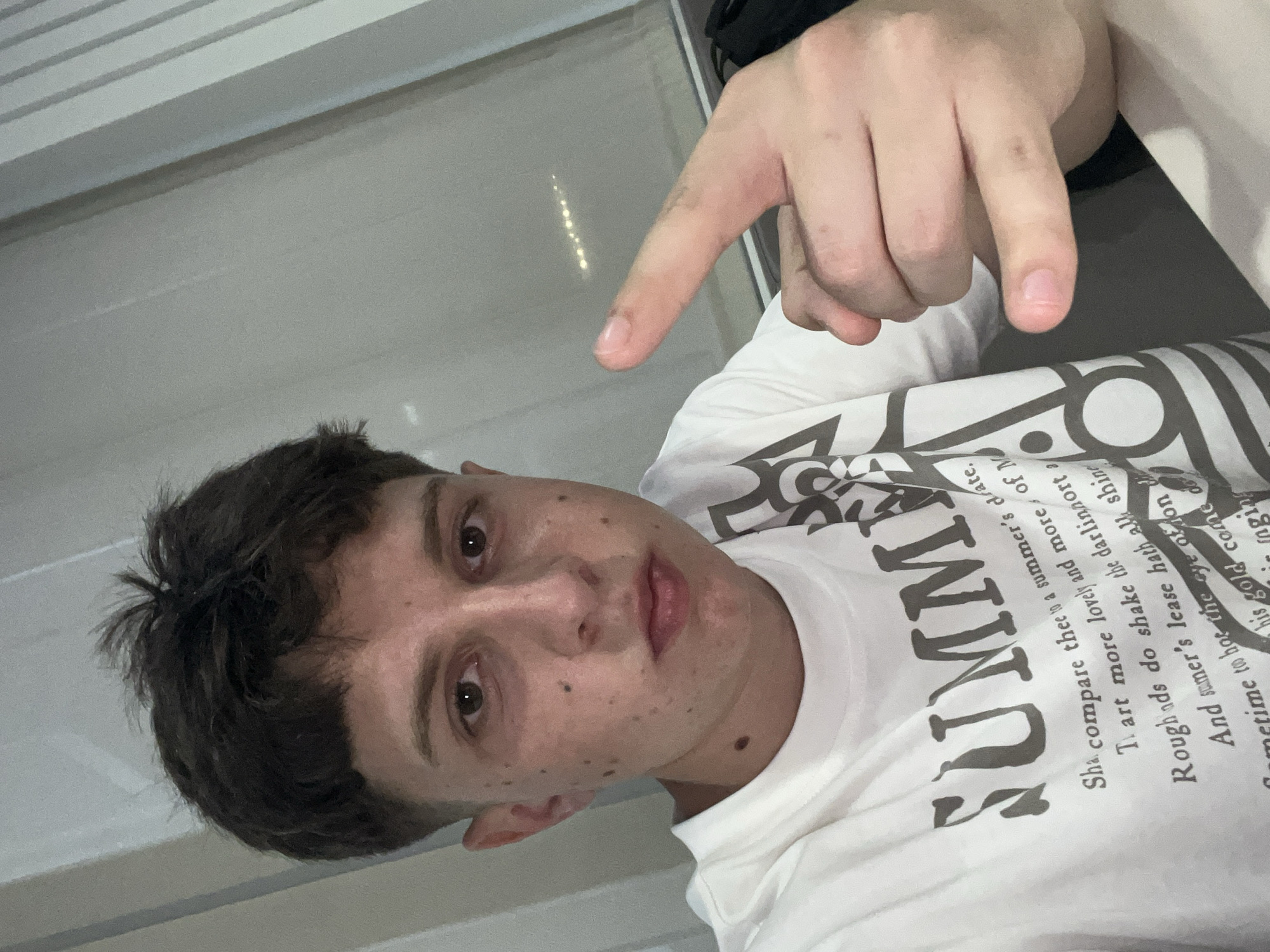 A close-up, rotated image shows a young man with short, dark hair, facing the camera and pointing with his right index finger toward the upper left corner of the frame. The man has fair skin with some freckles or spots visible on his face. He is wearing a white t-shirt with a design that includes the word "SUMMER" in large, bold letters and smaller text printed around it. The background appears to be a blurry, neutral-toned surface, possibly a wall or window. The lighting is somewhat diffused, creating a soft effect on the subject. No specific location in Jesolo, Italy, is evident in the image.