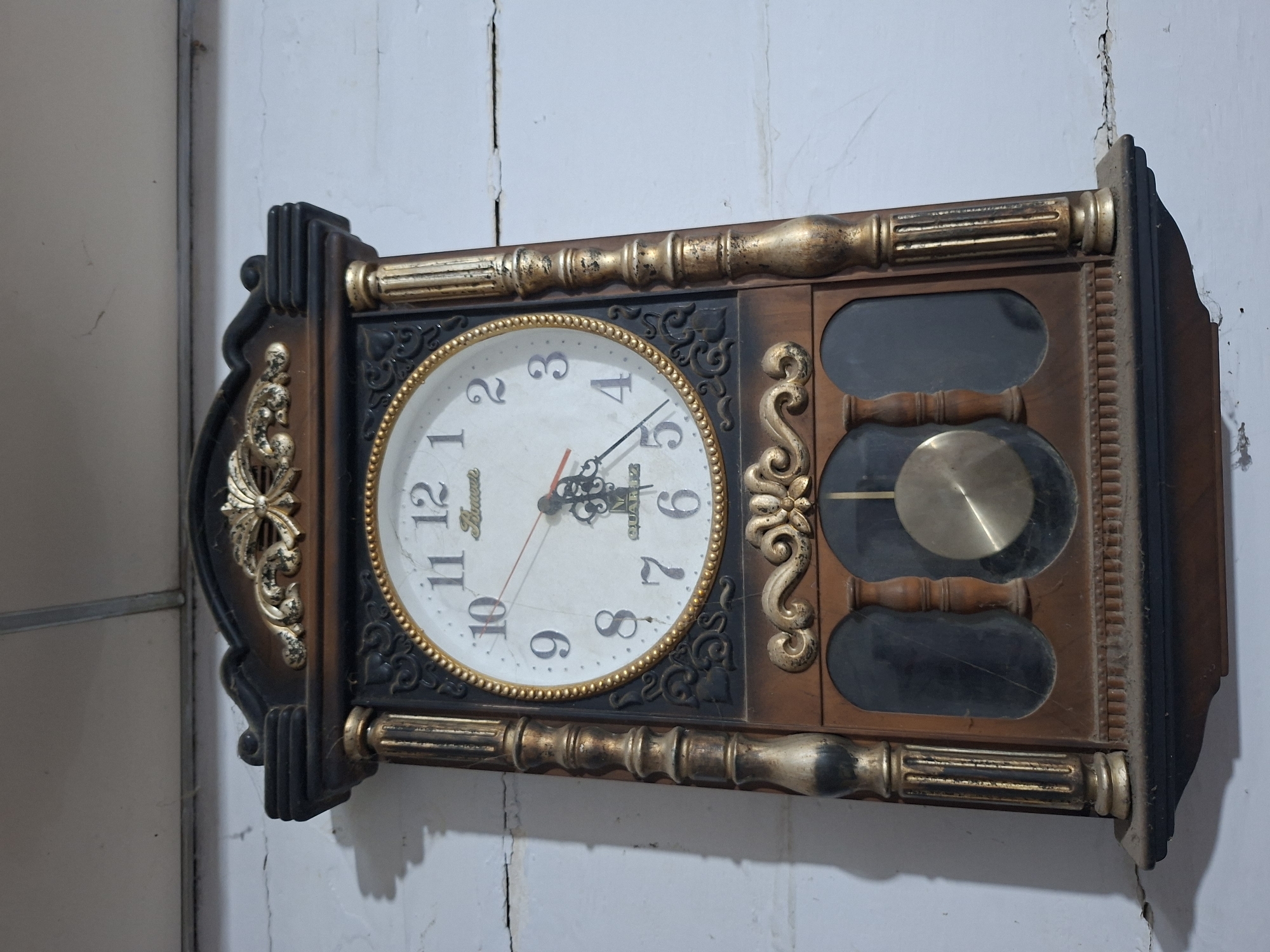 A vintage-style wall clock, designed as a pendulum clock, is centrally depicted, mounted on a light-colored, cracked wall. The clock features an ornate, rectangular wooden-look frame in dark brown and black, adorned with numerous gold-painted decorative elements, including fluted columns along the top and bottom, and intricate scrollwork and flourishes on the sides and below the clock face.
The main clock face is round and white, encircled by a gold beaded bezel. It displays black Arabic numerals from 1 to 12. The clock hands, comprising a short black hour hand, a longer black minute hand, and a thin red second hand, indicate the time is approximately 5:35. Visible text on the clock face includes "Hawa" above the "12" and "QUARTZ CHIME" above the "6" mark, suggesting the brand and features.
Below the clock face, a brass-colored pendulum bob is visible through a rectangular, transparent panel. Further below, two more dark, rectangular compartments, also behind transparent panels, are present. The wall behind and around the clock shows significant wear, characterized by multiple prominent vertical and diagonal cracks and some flaking paint, suggesting an aged or weathered interior environment. Dust and grime are noticeable on the top and right edges of the clock's frame. The scene is static, portraying an indoor setting in Lhokseumawe, Indonesia, with no specific activity taking place.