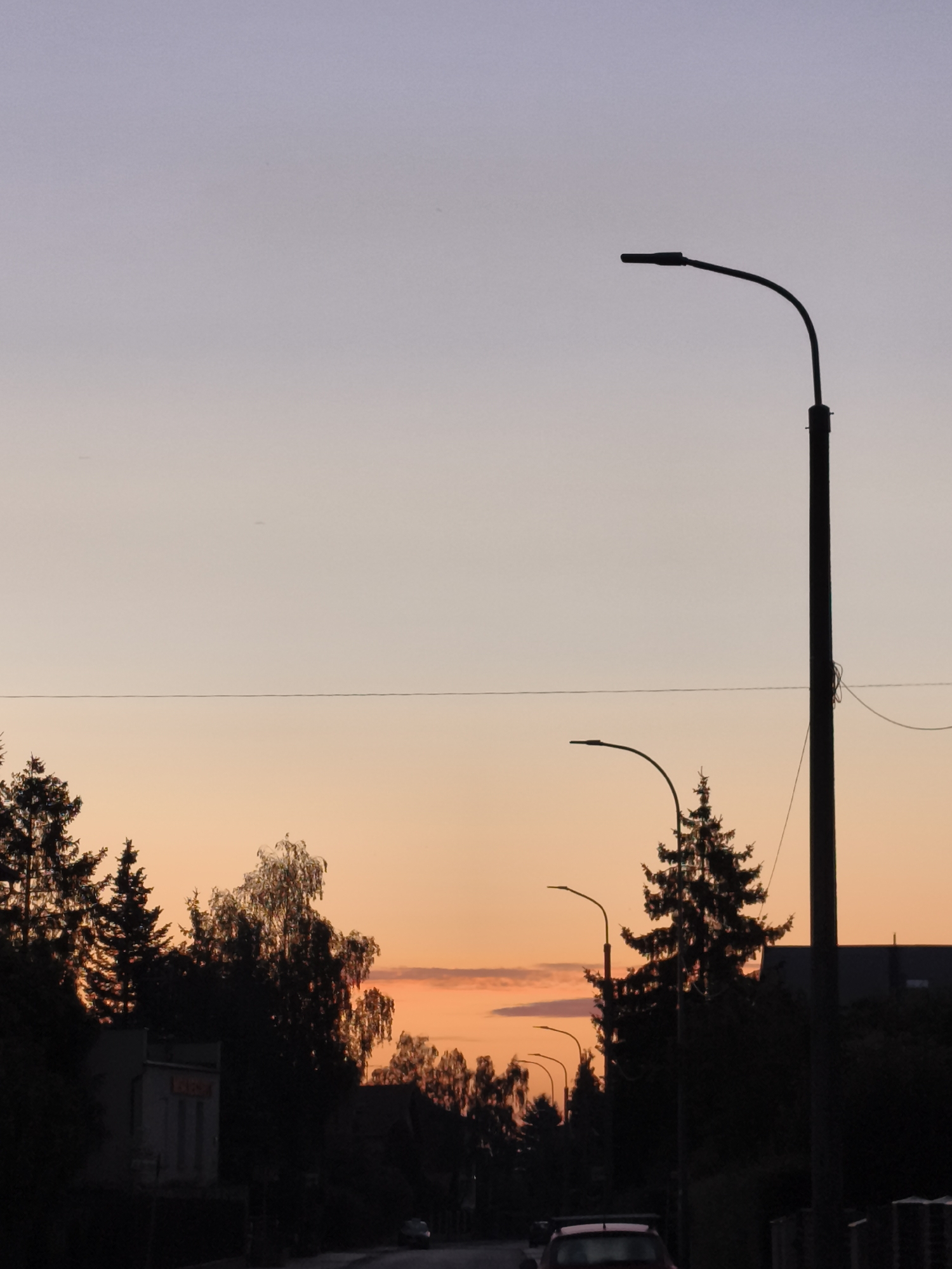 The image depicts a street scene at dusk in the city of Gdańsk, Poland. The sky is a gradient of pale lavender at the top fading into warm orange and pink hues near the horizon, suggesting the setting sun. Silhouetted against the sky are several streetlights, some with tall poles and curved arms, and the dark shapes of trees, including prominent evergreen trees and deciduous trees with sparse foliage. A thin, dark horizontal line, likely a power line or cable, stretches across the frame, dividing the sky.
In the foreground, a road is visible, leading into the distance. A dark-colored car with a spoiler is parked or driving on the road. To the left of the road, a low-rise building with a muted facade is partially visible, with some indistinct signage. The overall atmosphere is tranquil and somewhat moody due to the low light conditions and the silhouettes of the objects. The location in Gdańsk is not specifically identified within the image itself.