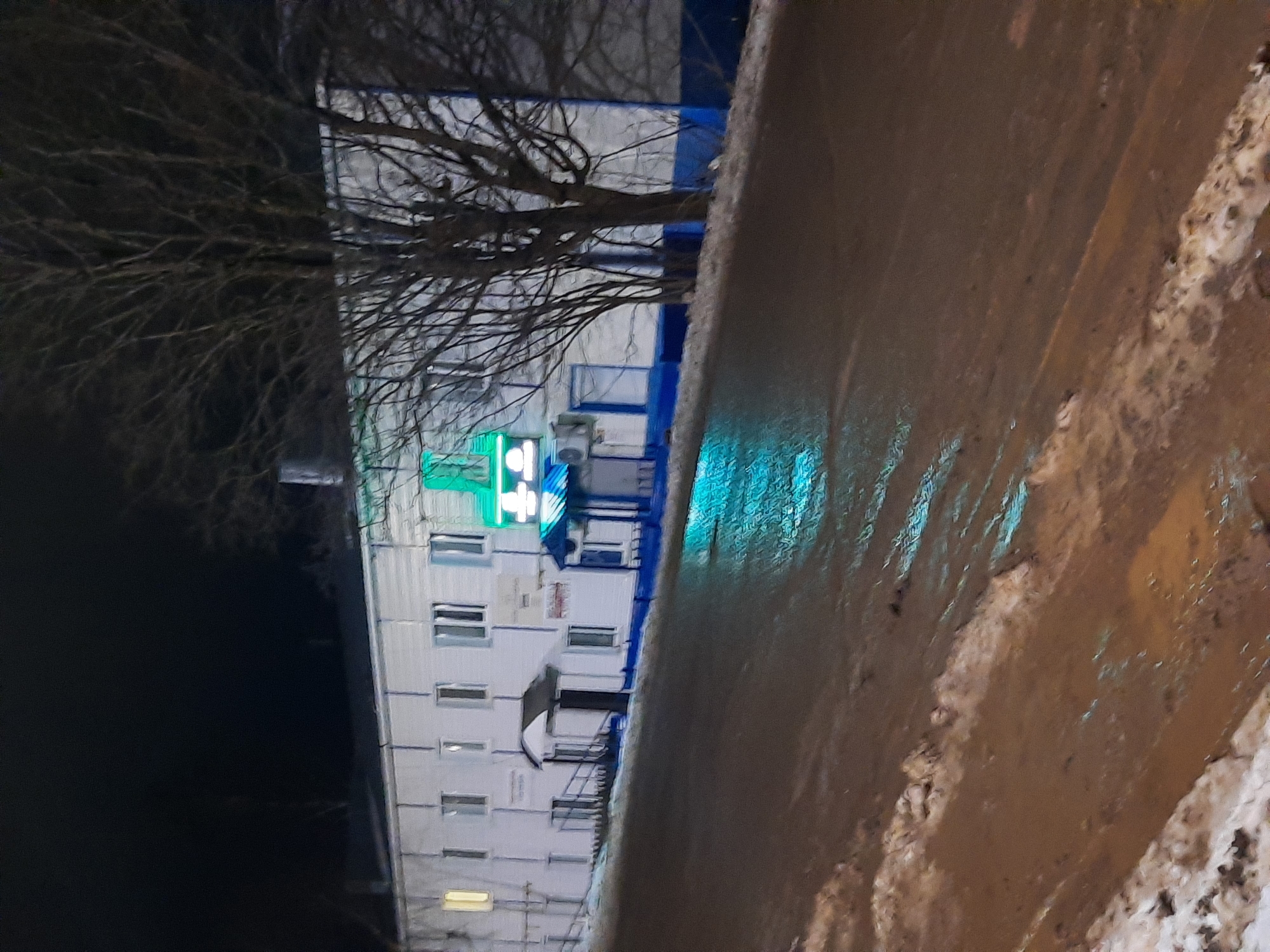 The image shows a multi-story building in Krasnogorskoye, Russia at night. A bare tree stands in front of the building. The building is white with blue trim, and a green neon cross indicates a pharmacy is located within. The road adjacent to the building is wet, and there is snow on the roadside. There are no people visible.