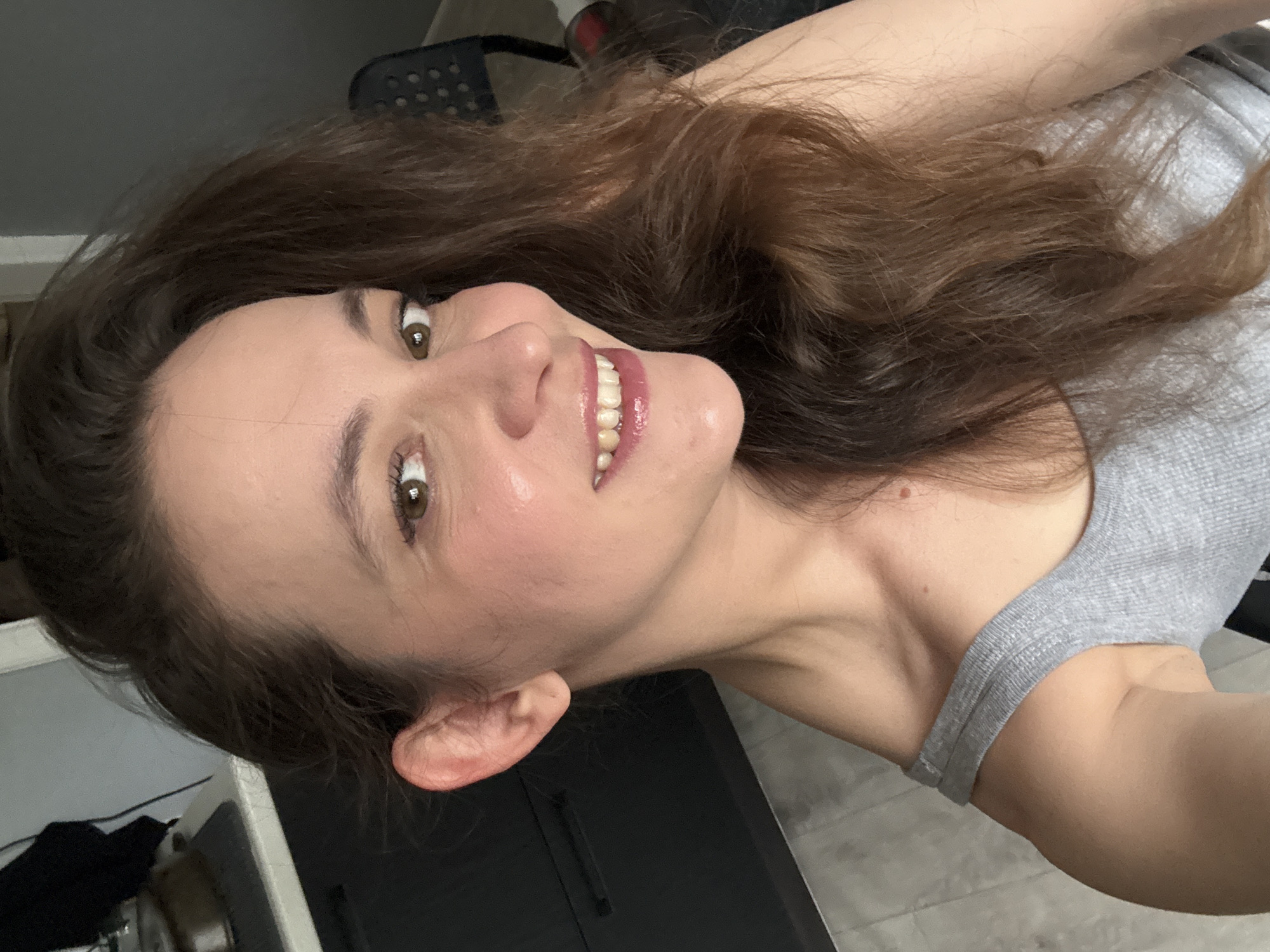 A medium-shot, eye-level selfie shows a woman with long, dark brown hair smiling and looking towards the camera. She is wearing a gray tank top. The background includes a dark cabinet and a light-colored floor, suggesting an indoor setting, possibly a home or a studio. There is no visible indication of the city of Chaikovsky, Russia.