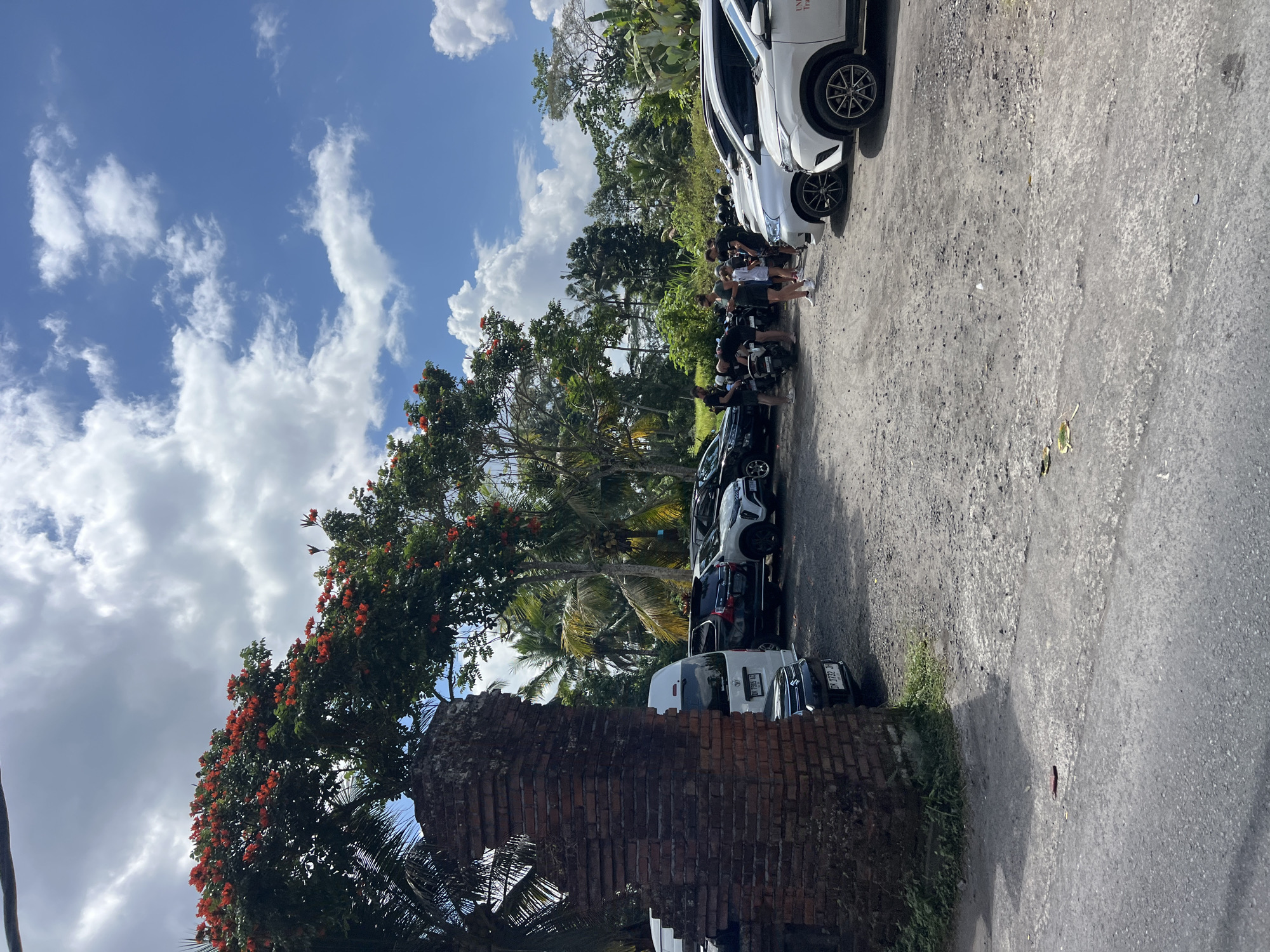 A group of people are gathered in a parking lot filled with vehicles, including cars and motorcycles. The lot is unpaved and appears to be in a tropical environment, with lush green foliage and palm trees in the background. The sky is blue with scattered white clouds. A partially constructed brick wall stands to the left of the frame, partially obscuring a white van. Some of the people are looking at their phones, while others appear to be interacting with each other or the motorcycles. The scene suggests a stop or gathering point for tourists or travelers in Ubud District, Indonesia, likely for an excursion or activity.