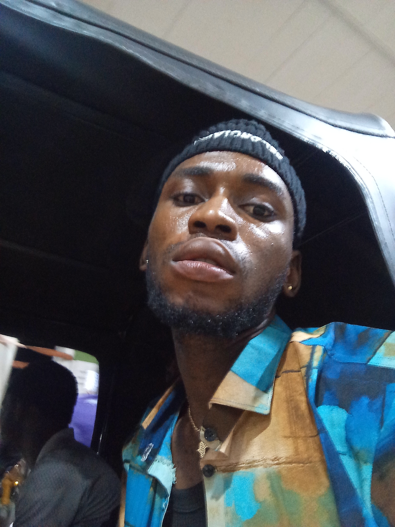 The image is a selfie taken inside a vehicle in Lokoja, Nigeria. The subject is a young Black male wearing a black beanie and a colorful short-sleeved button-up shirt with abstract blue, brown, and green patterns. He has a short beard and a gold stud earring in his right ear. He is looking directly at the camera, and his skin appears to be sweating. The background of the image shows the interior of a vehicle with dark-colored seats and a glimpse of what appears to be another person in the back seat on the left. The lighting suggests it is daytime, possibly indoors or under cover.