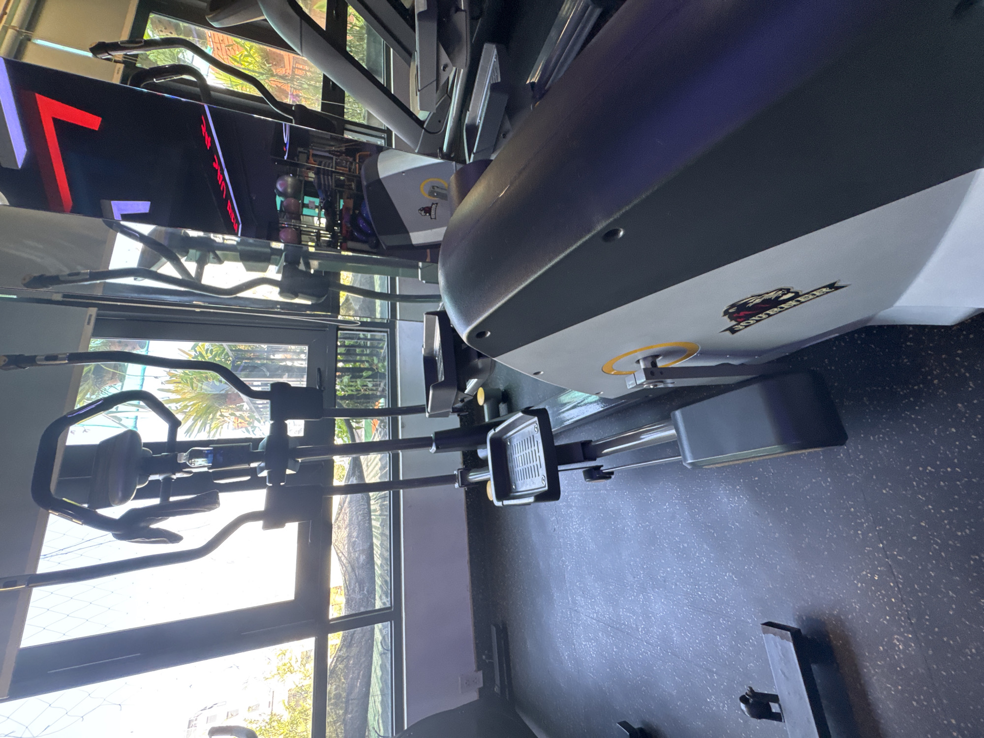 The image shows an indoor gym with exercise equipment. In the foreground, an elliptical machine is visible from the side, featuring a dark grey and white casing with a yellow circular accent and a logo that appears to read "JOURNEY." To the left, another elliptical machine is partially visible, with its pedals and handles. The background reveals large windows looking out onto an outdoor area with greenery and buildings, suggesting an urban setting. The gym floor is a dark, textured material. A large flat-screen television displaying a red and white graphic is mounted on the wall to the right. Other exercise equipment, including what looks like a treadmill, is also present in the background. The lighting appears to be a mix of natural light from the windows and artificial gym lighting. There are no people visible in the image. This scene is located in the city of Bac Nha Trang Ward, Vietnam.