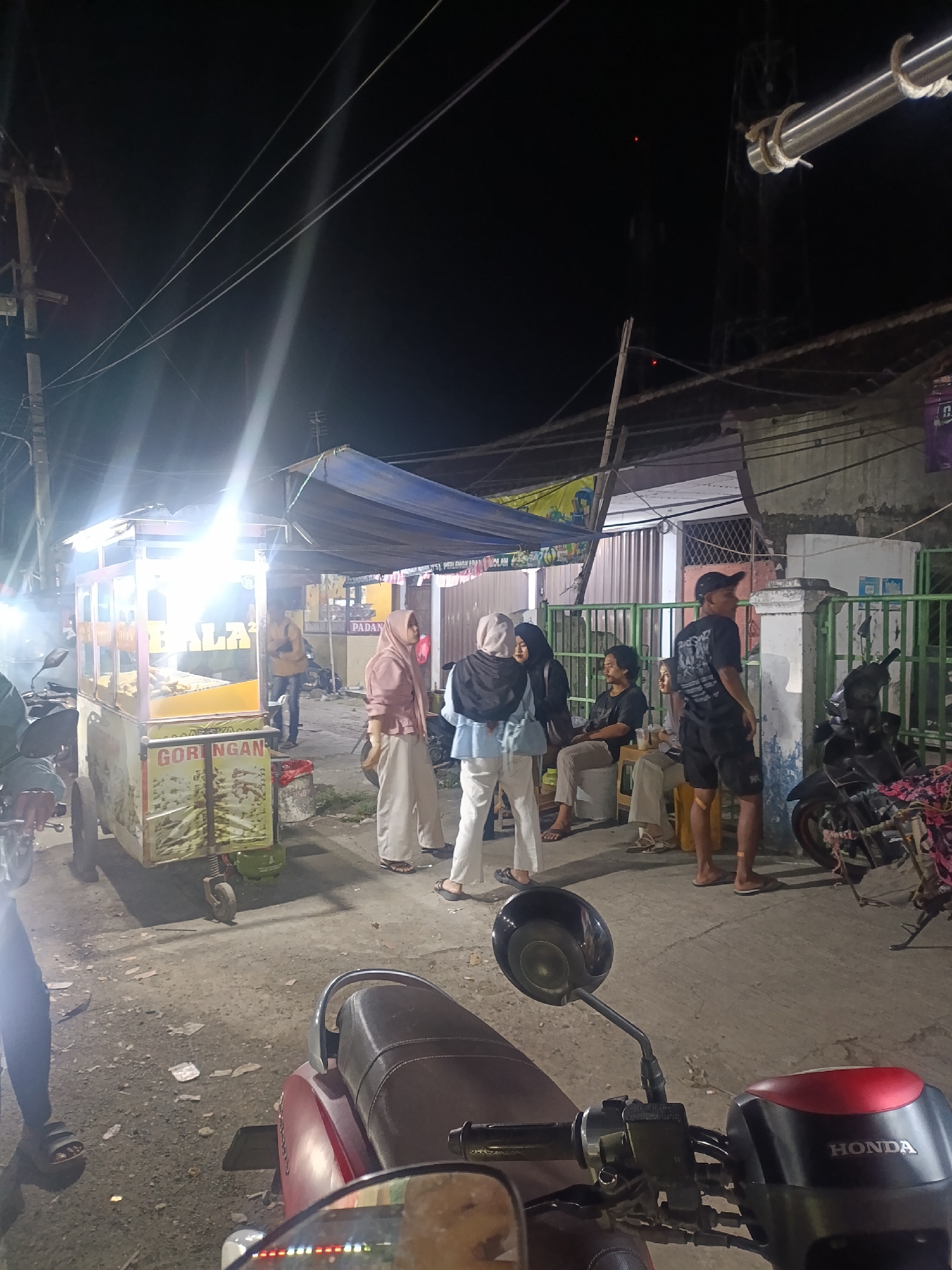 The image captures a street scene at night in Jonggol, Indonesia, likely near a food stall. In the foreground, the handlebars and part of the seat of a red Honda scooter are visible, suggesting the photographer is on a motorcycle. A food cart, brightly lit by overhead lights, is positioned in the middle ground. The cart displays the text "GORENGAN," indicating it sells fried food. Several people are gathered around the cart and in the background.

To the left of the cart, a person wearing a light pink hijab and matching pants stands, facing away from the camera. Next to them, two individuals, one in a light blue shirt and white pants and the other in a black hijab, are facing each other. Further back, a man in a black t-shirt and shorts stands and looks to the right, while a woman in a hijab sits on a stool, also looking towards the right. Another man in a dark t-shirt and lighter pants sits on a stool to their left.

In the background, a building with a green metal fence is visible, along with more electrical wires and poles, typical of an urban Indonesian setting. A motorcycle is parked near the fence. The ground is paved and appears somewhat dusty. The overall lighting suggests it is nighttime, with artificial lights from the food cart and other sources illuminating the scene. The atmosphere appears casual, with people congregating around the food stall.