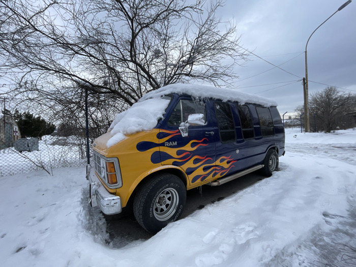 A customized Dodge Ram van is parked outdoors in a snow-covered environment, likely a suburban or semi-industrial area of Belgrade, Serbia, under an overcast sky. The van features a yellow hood and front quarter panels with prominent orange and red flame decals that transition into a dark blue body, extending to the rear. A layer of snow covers the van's roof and portions of the hood. The vehicle is equipped with dark tinted side windows, chrome side mirrors, a chrome front bumper, and chrome five-spoke wheels fitted with Goodrich Radial T/A tires. A chrome running board is visible along the side. The ground surrounding the van is blanketed in white snow, revealing tire tracks and footprints. To the left, a chain-link fence and a bare deciduous tree with intricate branches are visible, with a small, weathered structure and evergreen shrubs beyond the fence. To the right, a tall utility pole with a streetlamp extends skyward, supporting overhead power lines.