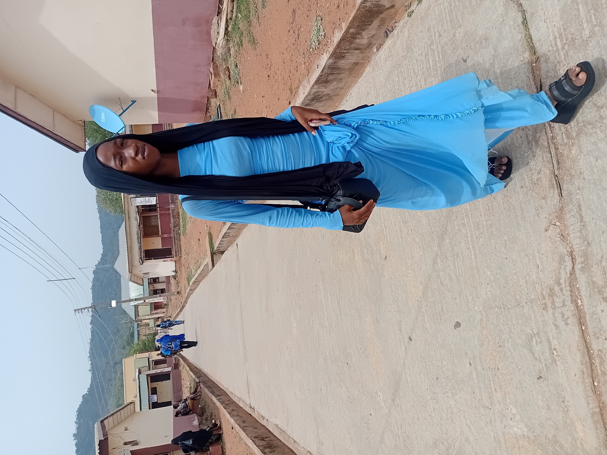 The image shows a woman wearing a long blue dress and a black headscarf, standing on a paved road in Lokoja, Nigeria. She is wearing black sandals and holding a black bag or purse. A concrete drainage ditch runs alongside the road. In the background, there are buildings, a mountain range, and overhead power lines. Other people are visible in the distance. A satellite dish is affixed to one of the buildings. The image appears to be taken during the daytime.