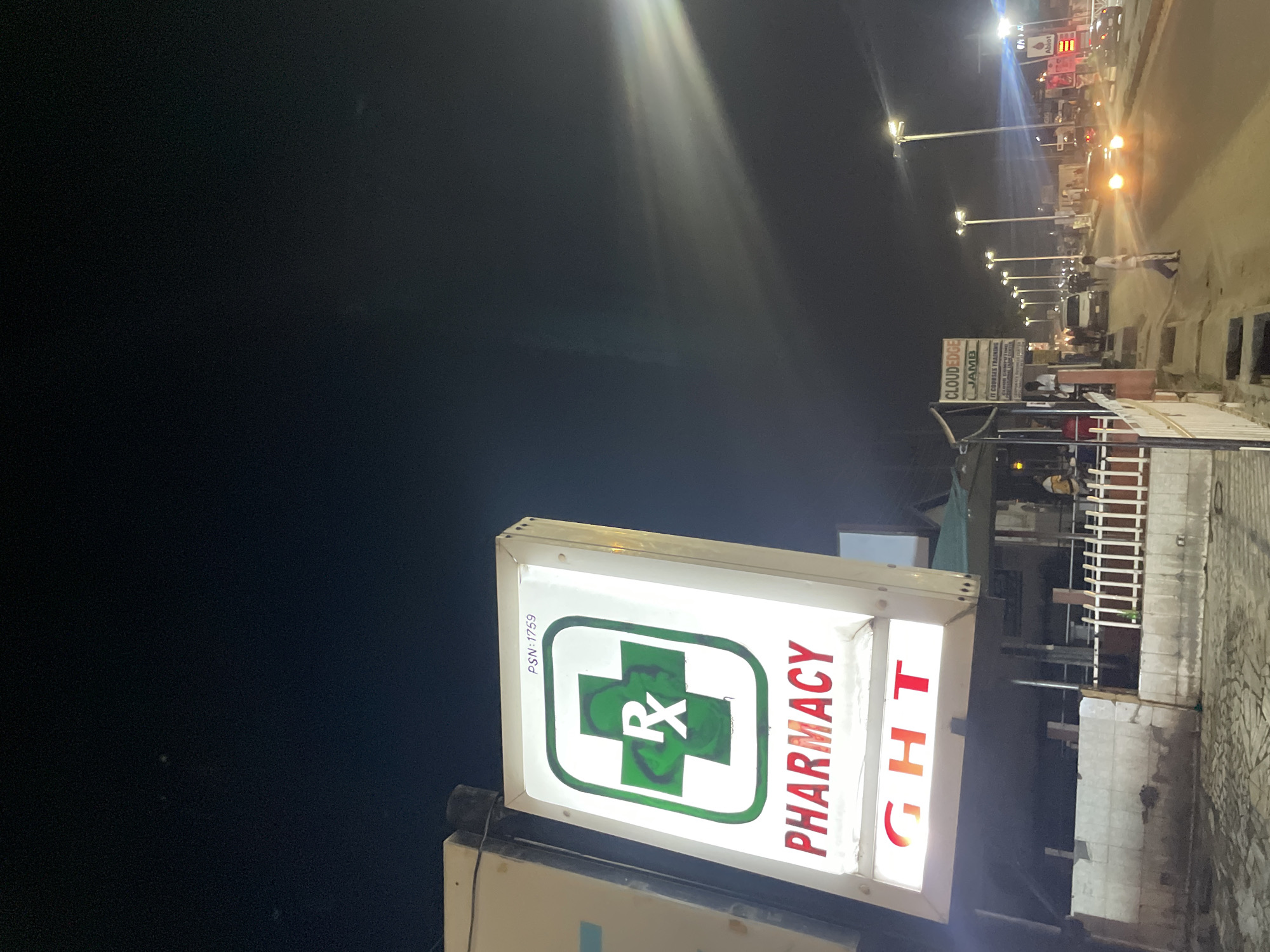A nighttime photograph shows a lit pharmacy sign on a street in Ado, Nigeria. The sign is rectangular and features a green cross with the letter "R" inside it, along with the word "PHARMACY" in red, followed by the letters "GHT." The sign is illuminated and casts light on the surrounding area. To the right of the pharmacy sign, there is a sidewalk and a metal fence. Further down the street, there are other lit signs and streetlights, as well as a few people walking. The overall impression is that the photo was taken at night in an urban environment.