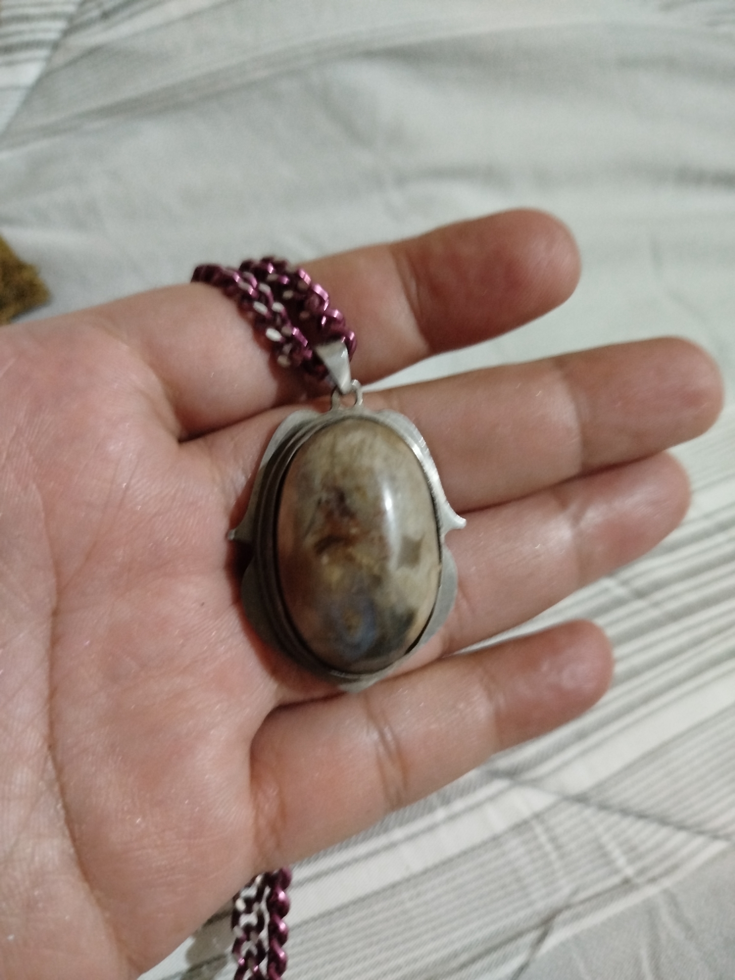 A hand holds a silver pendant with an oval, polished stone featuring natural swirling patterns of light brown, grey, and blue. The pendant is suspended from a metallic chain with alternating dark purple and silver links. The background appears to be a striped fabric, possibly a blanket or bedding, with alternating light and dark grey stripes. The scene is indoors, with soft lighting that highlights the textures of the hand and the pendant. There is no discernible information about the location in Bandungrejo, Indonesia.