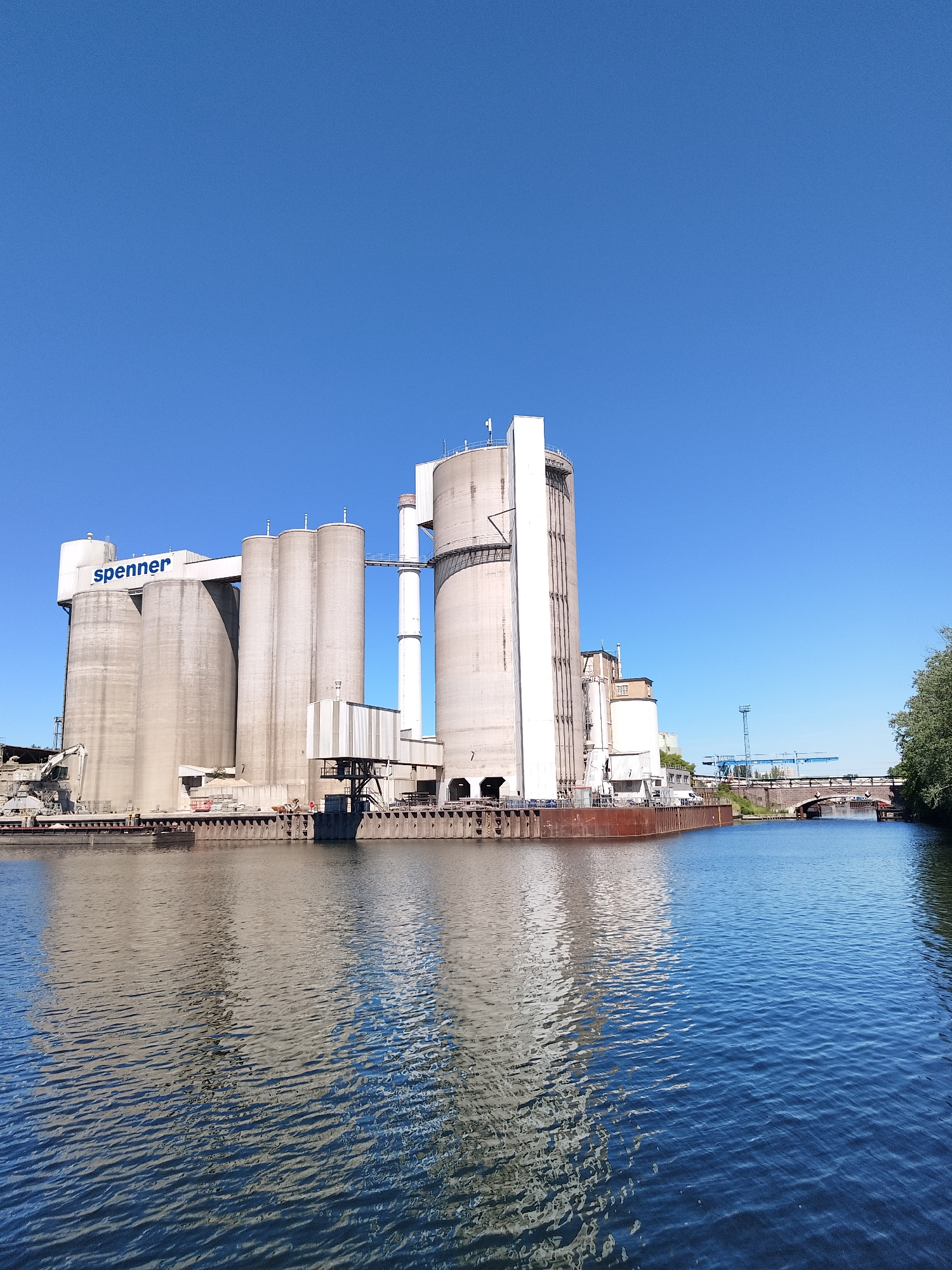 The image depicts a large industrial complex, possibly a grain elevator or a similar processing facility, situated along a waterway, likely a river or canal. The complex comprises a cluster of tall, cylindrical concrete structures, some marked with the name "spenner" in blue lettering. A network of walkways and conveyor systems connects the different structures. The complex is built on a platform or barge extending into the water, with various piers, support structures, and machinery visible.
In the background, a bridge spans the waterway, and a crane stands alongside it. The sky is clear and blue, suggesting a sunny day. The water surface reflects the structures and the sky, creating a mirrored effect. On the right side of the image, trees partially obscure the view. The location is Unknown, Unknown.