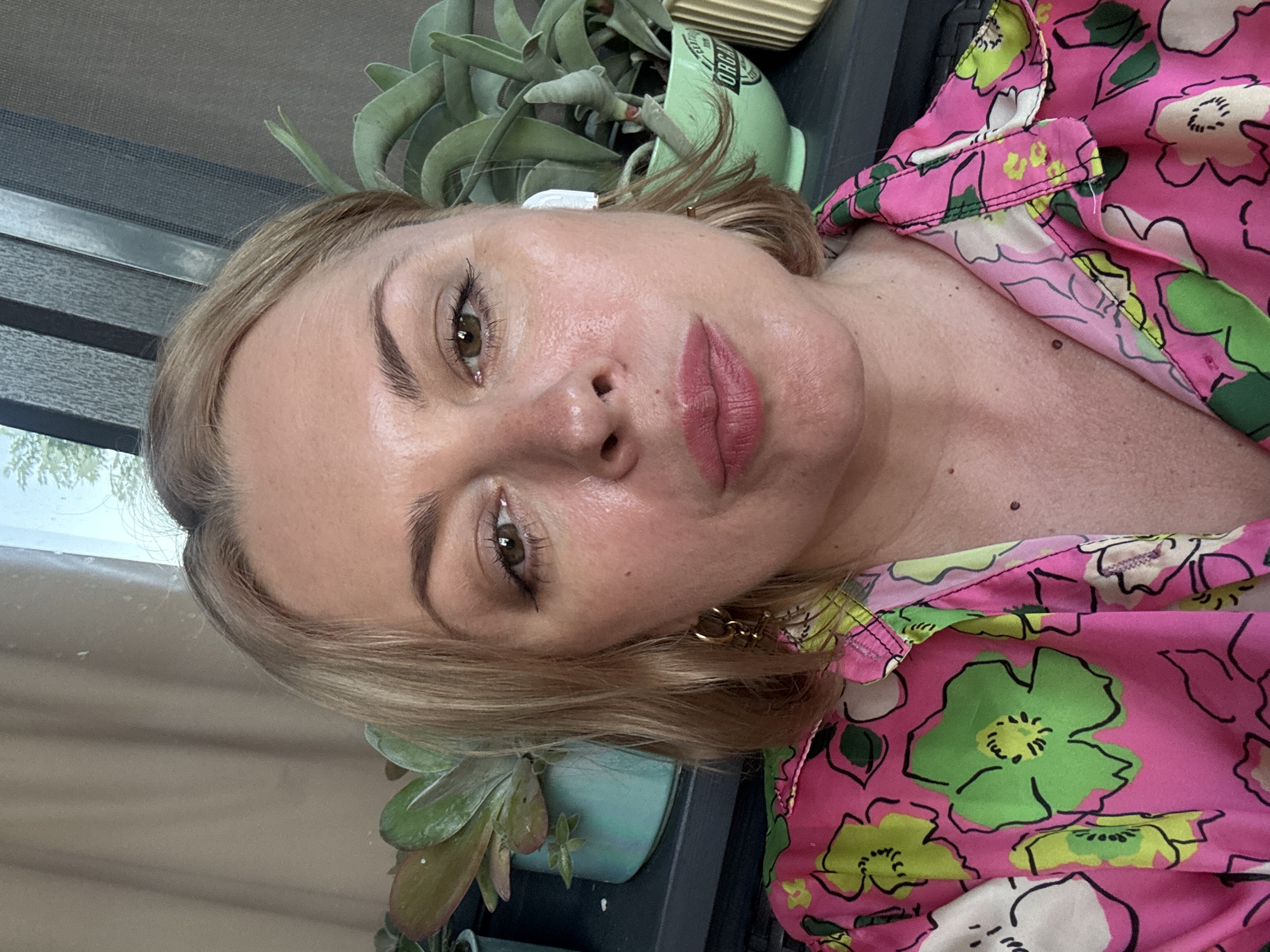 A close-up, waist-up, outdoor selfie shows a light-skinned woman with blonde hair and fair skin. She is wearing a vibrant pink shirt with a floral pattern in green and white. She has on gold hoop earrings and a white wireless earbud in her right ear. Her makeup includes pink lipstick and eyeliner. Behind her, there are plants in light green and teal pots, and a window with blinds. The image does not contain any identifiable landmarks or features that would indicate the location as Sochi, Russia.