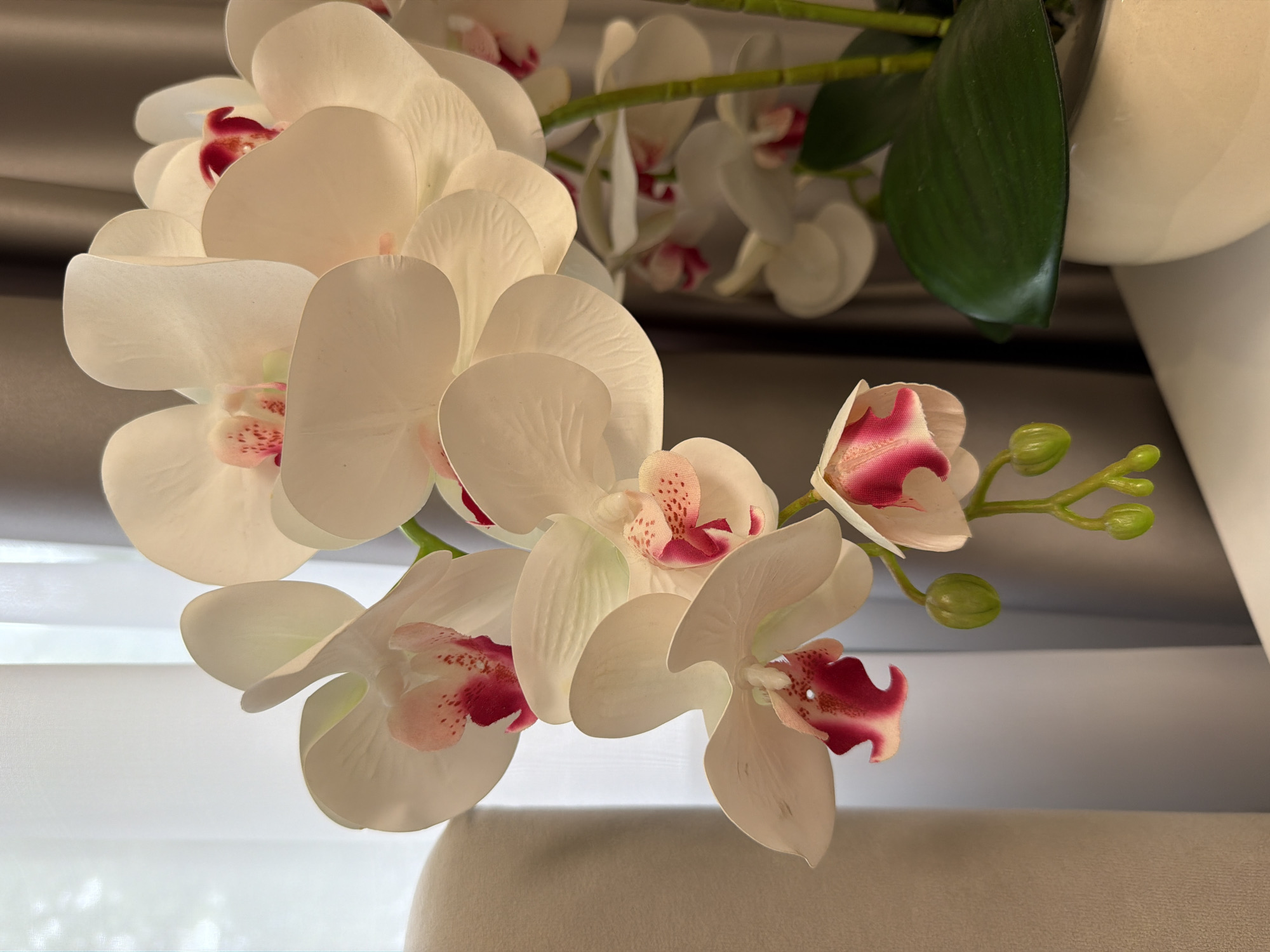 The image features a close-up of a cluster of artificial white orchids with pink and red centers, some with visible buds. The orchids are arranged on a green stem and appear to be part of a larger floral arrangement. The background is softly blurred, showing draped curtains in a neutral tone and hints of a white vase. The lighting suggests it is daytime, possibly indoors with natural light filtering through. The overall impression is one of decorative elegance, likely in a domestic or commercial setting. There is no text visible in the image.