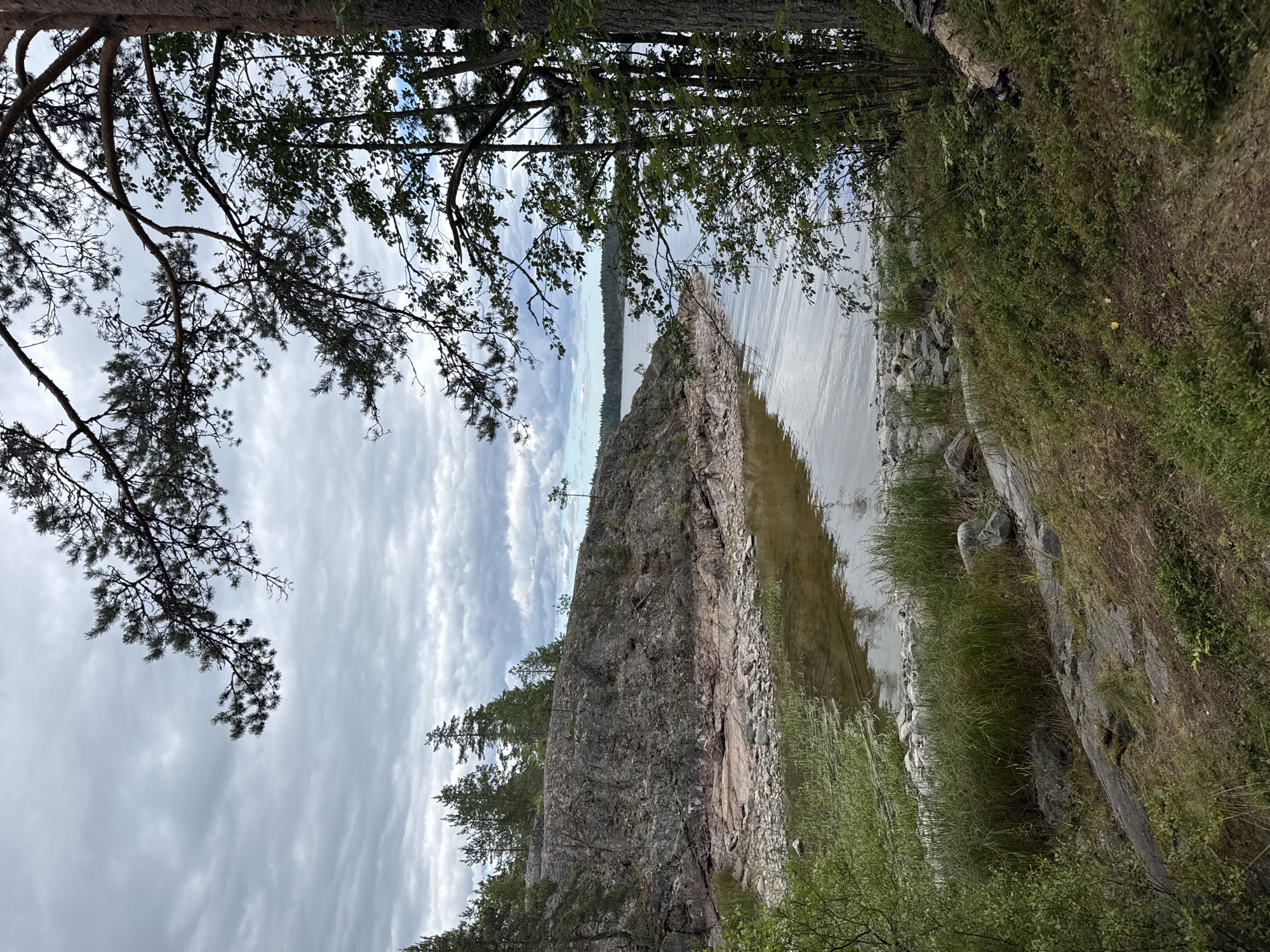 The image depicts a natural landscape characterized by a rocky shoreline and a body of water under a cloudy sky. In the foreground, dense green foliage and scattered rocks line the edge of the water. The water itself appears calm, with ripples reflecting the overcast sky. Beyond the immediate shore, a large, grey, rocky formation rises, sparsely populated with small trees and shrubs. The background reveals a distant horizon with more trees and a vast expanse of water. The overall scene suggests a remote, undeveloped natural environment. The image is taken from a slightly elevated position, looking out towards the water and the rocky outcrop. The lighting is diffused, consistent with an overcast day.