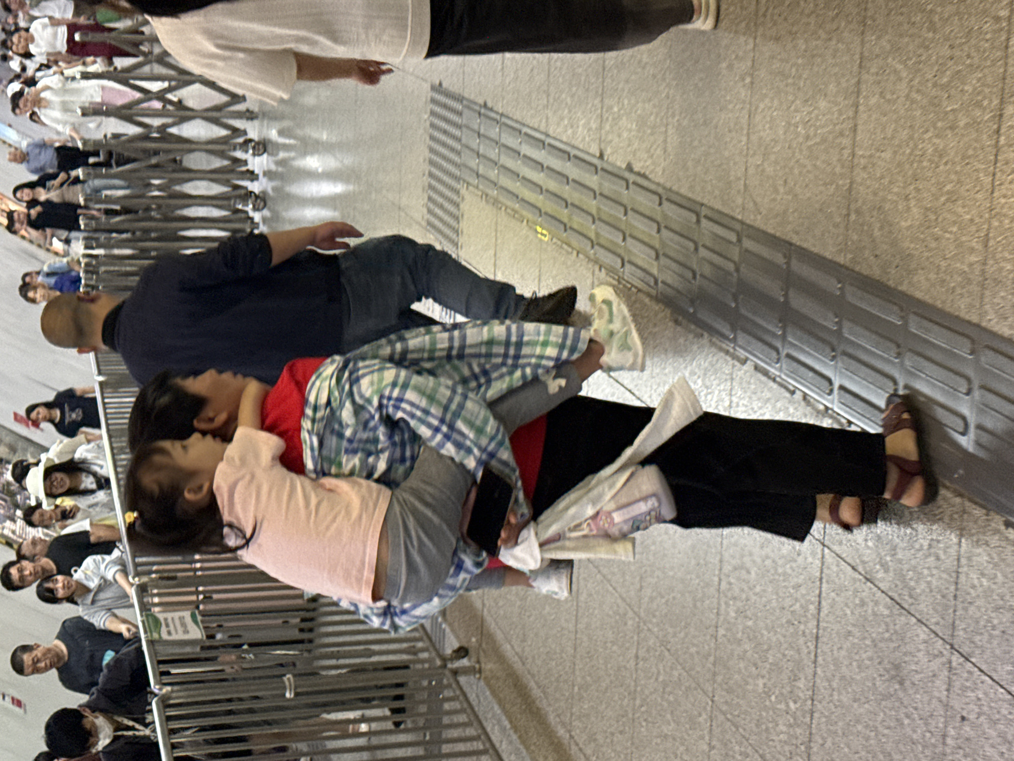 In the city of Yuzhong District, China, at street level, a man is carrying a young girl on his shoulders. The man, wearing a plaid shirt over a red shirt and dark pants, is walking forward. The girl, in a pink shirt and gray pants, is looking forward, her arm around the man's neck. Beside them, a woman in black pants and sandals is walking. In the background, various people are visible walking in the same direction. A metal accordion gate is partially open on the right side of the image. The environment appears to be an indoor walkway or metro station, with a tiled floor and a metal railing on the left.
