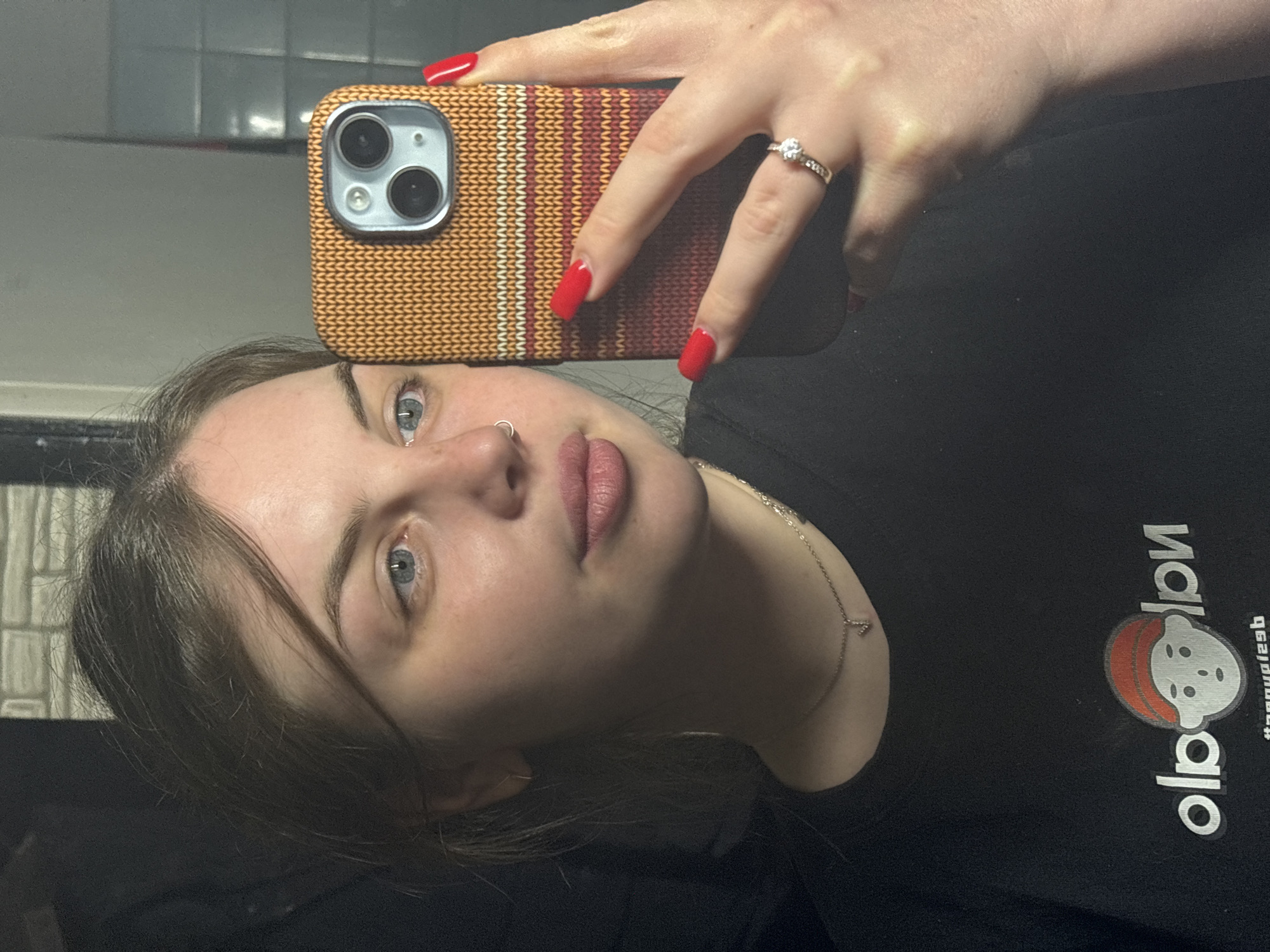 A young woman is taking a selfie. She is holding a smartphone with a textured orange and brown case in her right hand. The phone has a dual-lens camera system. Her nails are painted bright red, and she is wearing a ring on her ring finger. She has fair skin, light blue eyes, and a nose ring. She is wearing a black t-shirt with a logo that is mirrored in the image. The location is indoors, possibly a bathroom, with white tiled walls visible in the background. The image is slightly rotated. There is no evidence to suggest the location is in Новосельское сельское поселение, Ukraine.